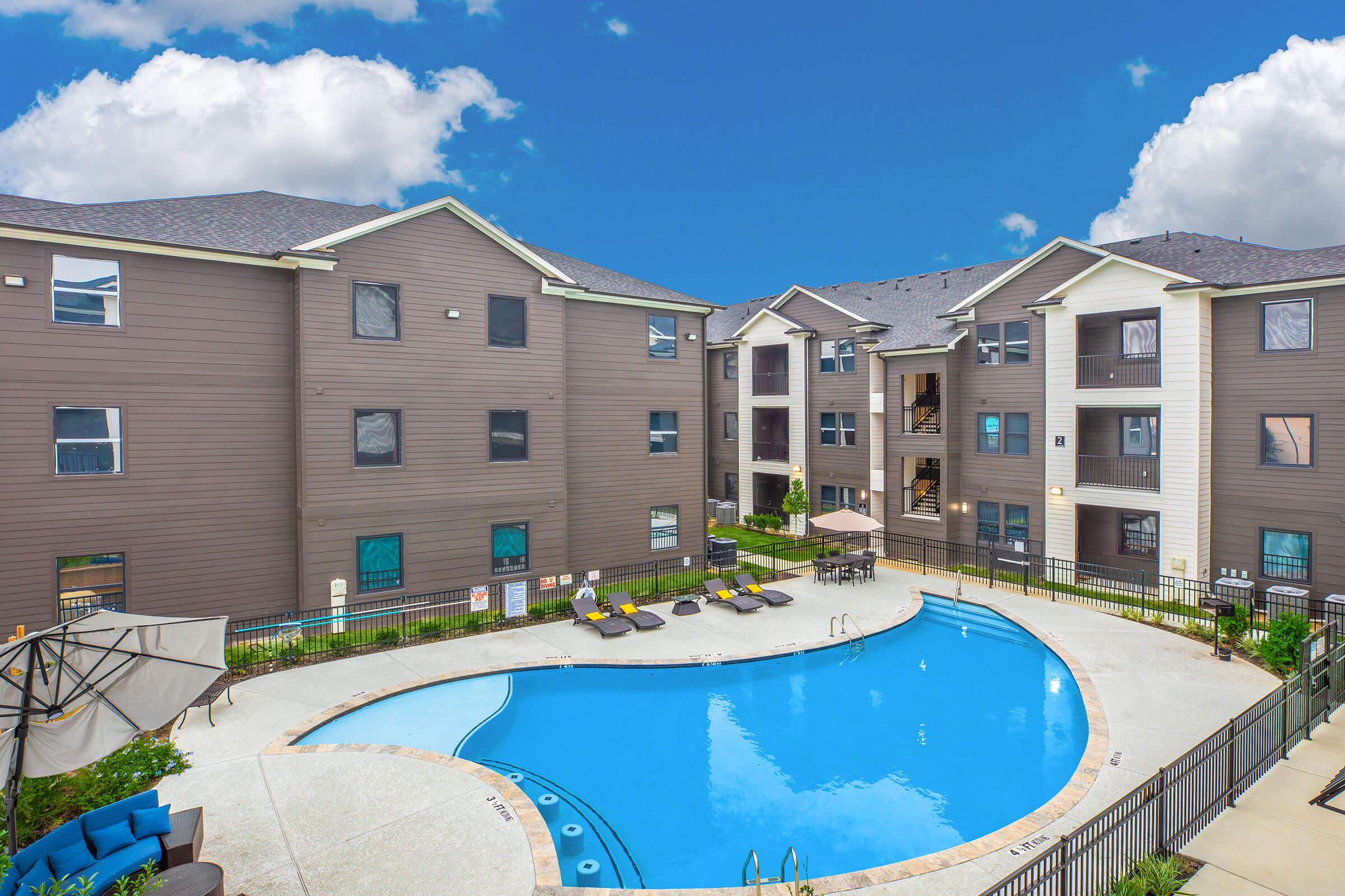 A view of a residential apartment complex featuring a swimming pool surrounded by lounge chairs. The buildings are modern, with a mix of dark and light exterior colors, and the sky is partly cloudy. Lush greenery is visible around the pool area, creating a relaxing outdoor environment.