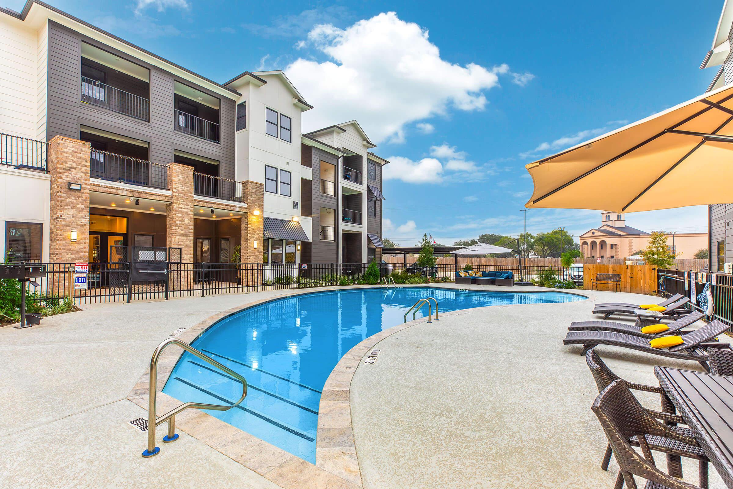 A scenic pool area featuring a curved swimming pool with clear blue water, surrounded by lounge chairs and an umbrella. In the background, a modern apartment building with multiple balconies and a bright sky overhead, creating a relaxing atmosphere for residents and visitors.