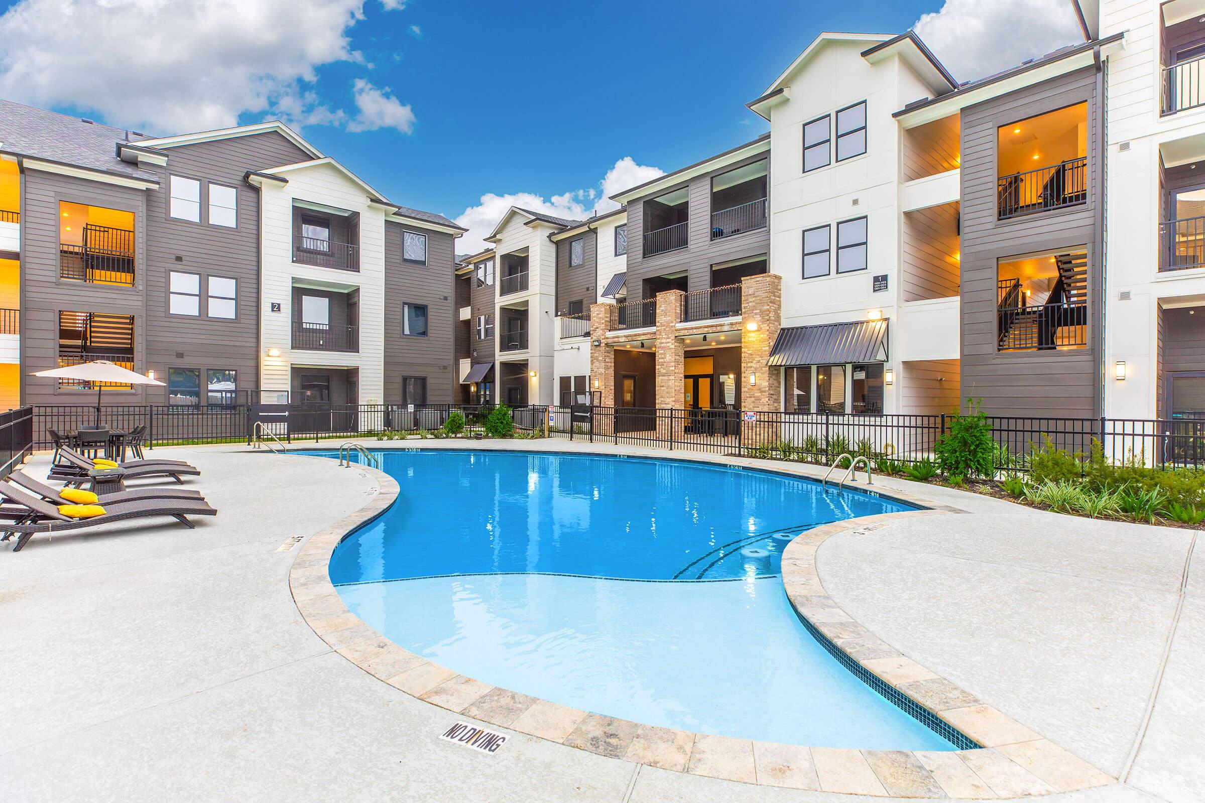 A serene apartment pool area featuring a large, inviting swimming pool surrounded by lounge chairs. Modern apartment buildings in the background, with balconies and outdoor seating. Lush greenery and a clear blue sky enhance the tranquil atmosphere.