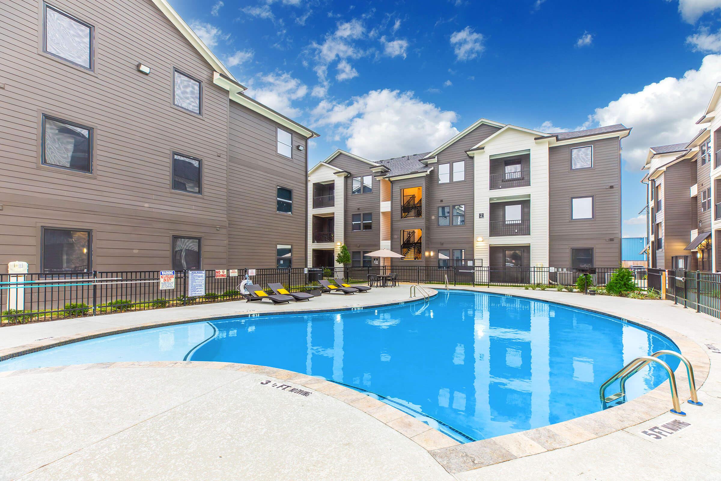 A modern apartment complex featuring a spacious swimming pool surrounded by lounge chairs. The architecture consists of multi-story buildings with a mix of brown siding and large windows. A bright blue sky with scattered clouds enhances the inviting atmosphere of the pool area.