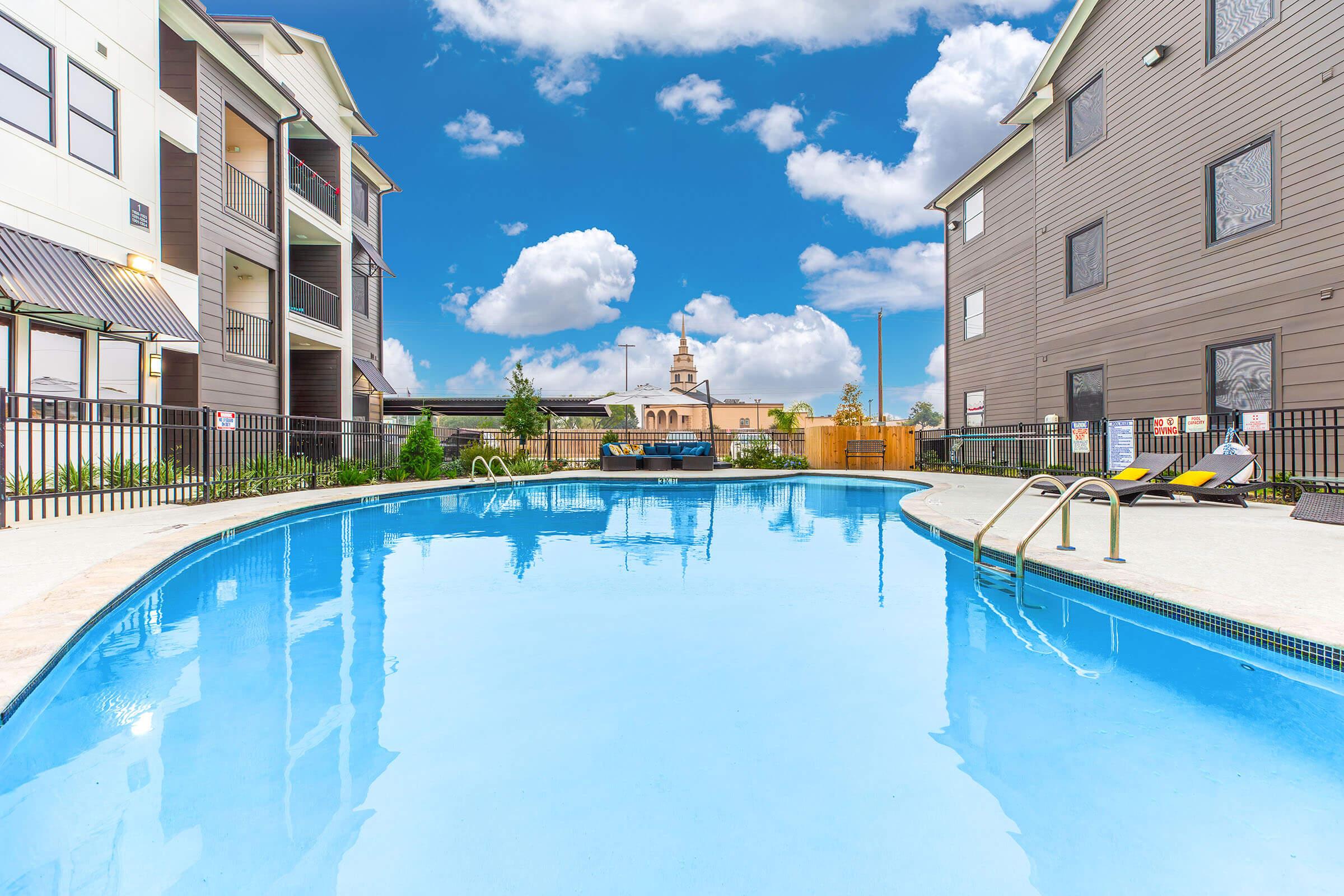A bright and inviting swimming pool surrounded by modern apartment buildings. The pool features a clear blue surface reflecting the sky and fluffy clouds. There are lounge chairs on the poolside, and greenery adds a touch of nature to the serene atmosphere.