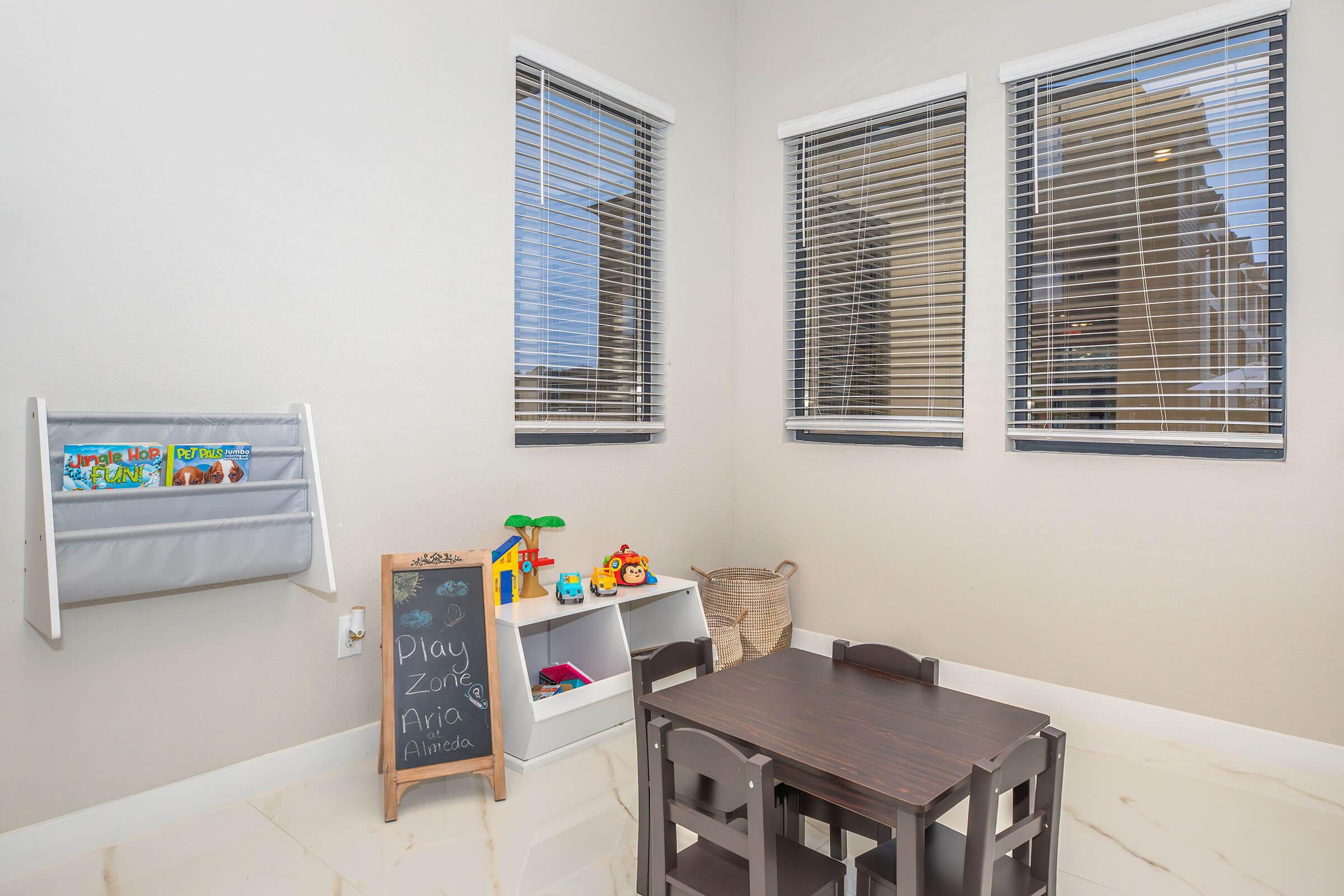 An interior playroom featuring a small black table and chairs for children, a white shelving unit with toys and books, a chalkboard labeled "Play Zone," and large windows with blinds allowing natural light to enter the space. The walls are painted a light color, creating a bright and inviting atmosphere.