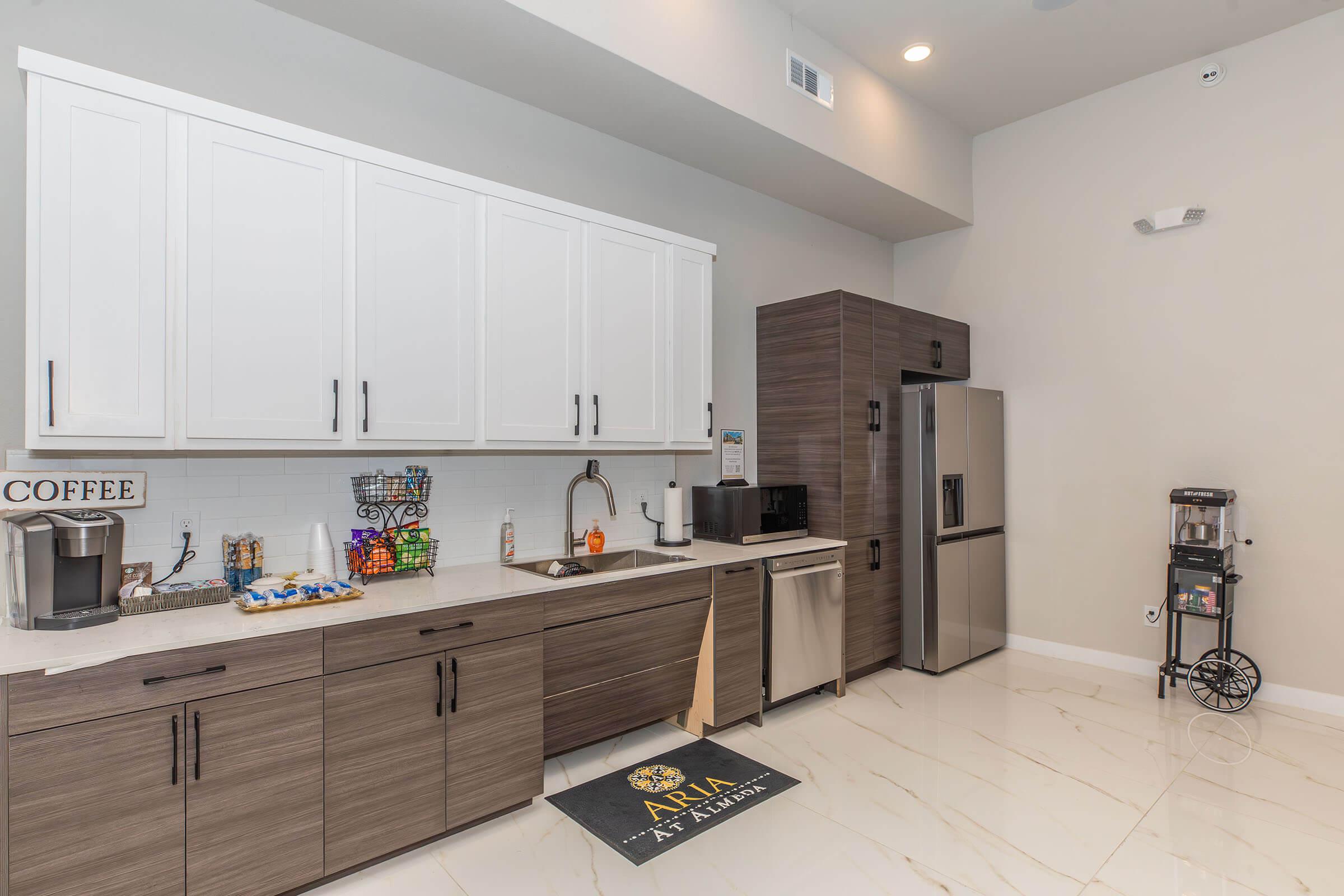 A modern kitchen featuring white and dark wood cabinetry, a stainless steel refrigerator, coffee machine, sink, and countertop. The space includes a candy display and a branded mat with "ARIA at Abilene" on the floor. Bright lighting enhances the clean, contemporary design of the area.