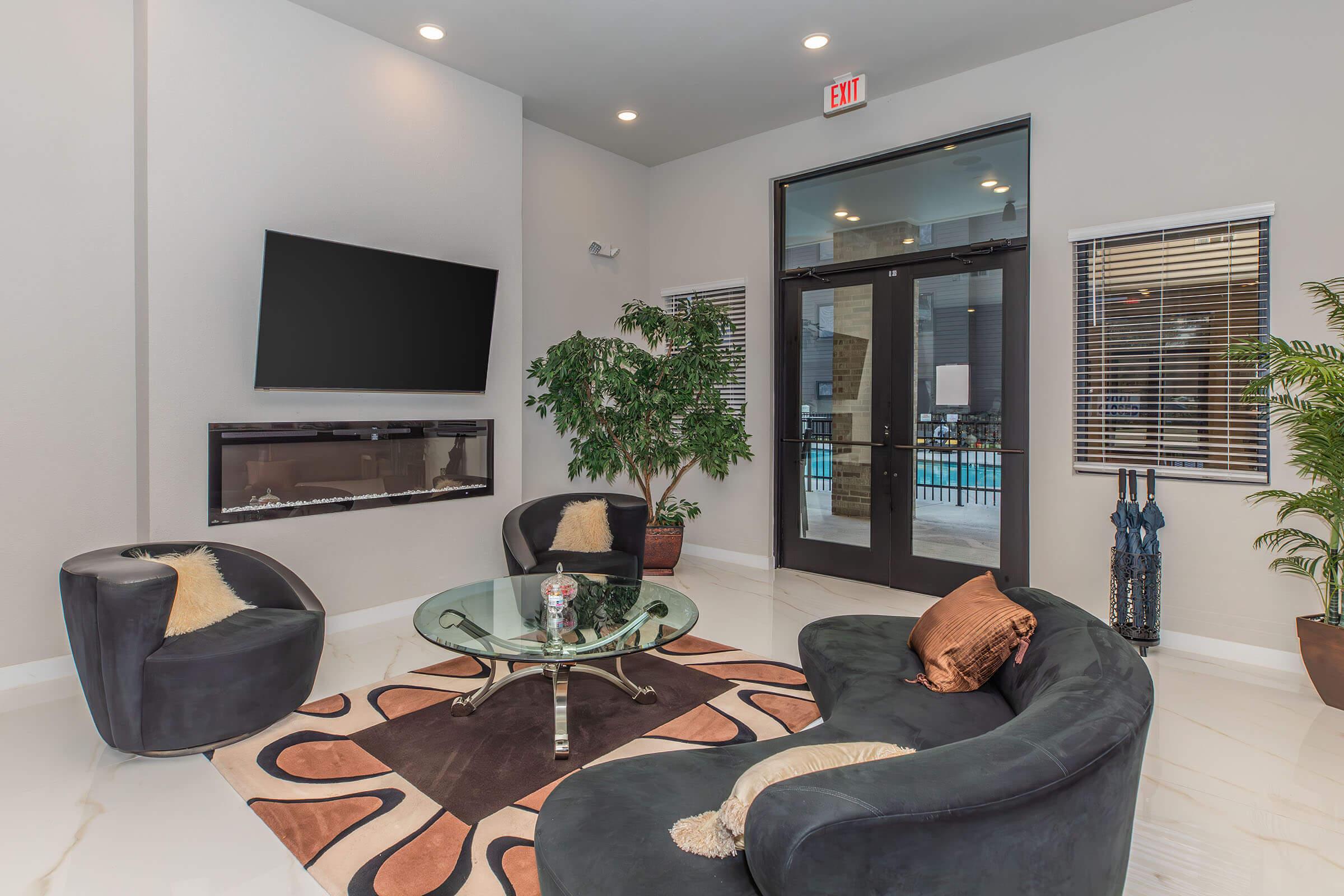A modern lounge area featuring two round black chairs with plush cushions, a glass coffee table on a decorative rug, and an elegant plant. A wall-mounted TV and large glass doors leading to an outdoor pool area complete the inviting atmosphere. Natural light streams in from the windows.