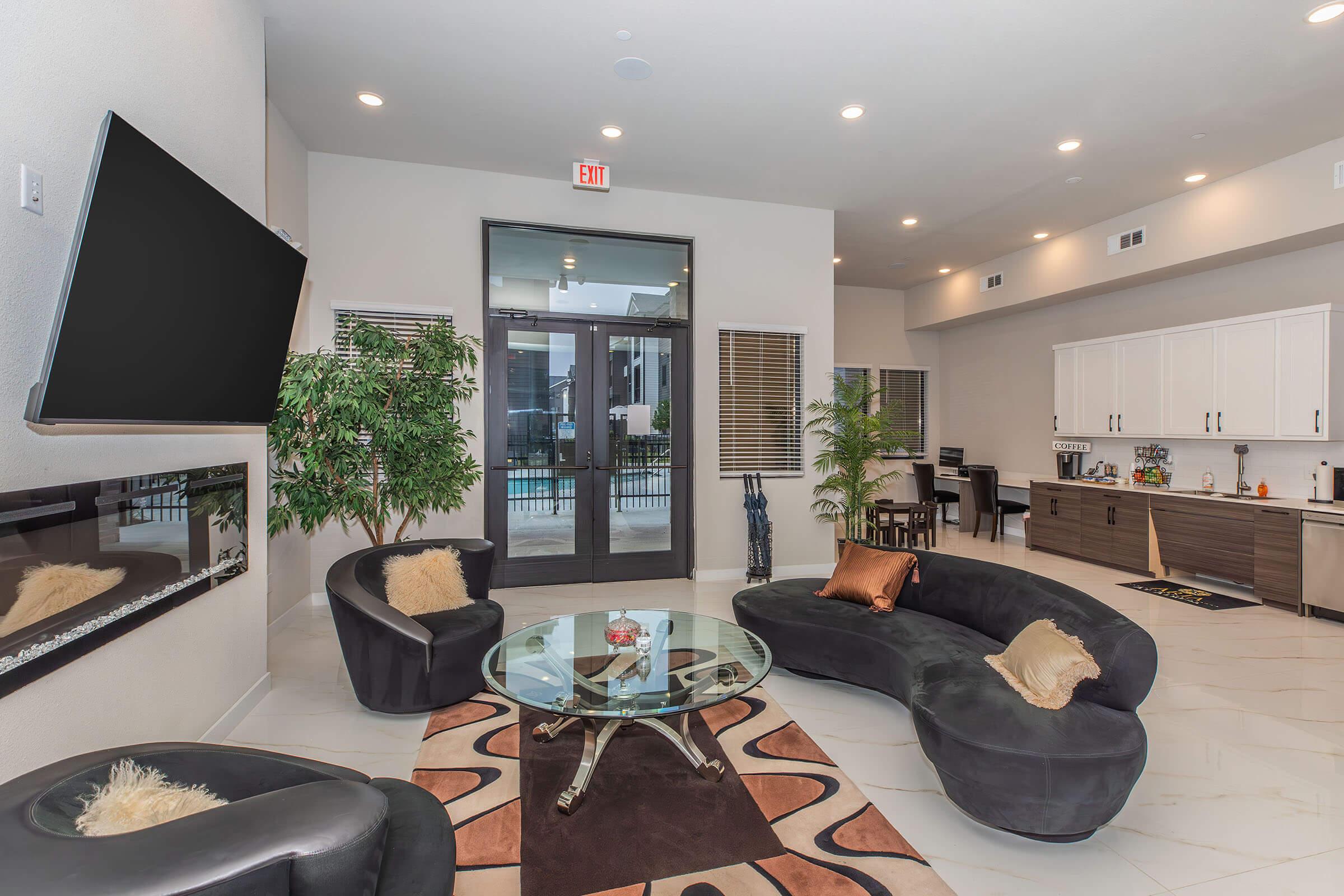 Modern lounge area featuring two curvy black sofas around a glass coffee table. In the background, there is a modern kitchen with white cabinetry and a dining area. Large windows provide natural light, and decorative plants add a touch of greenery. A large flat-screen TV is mounted on the wall.