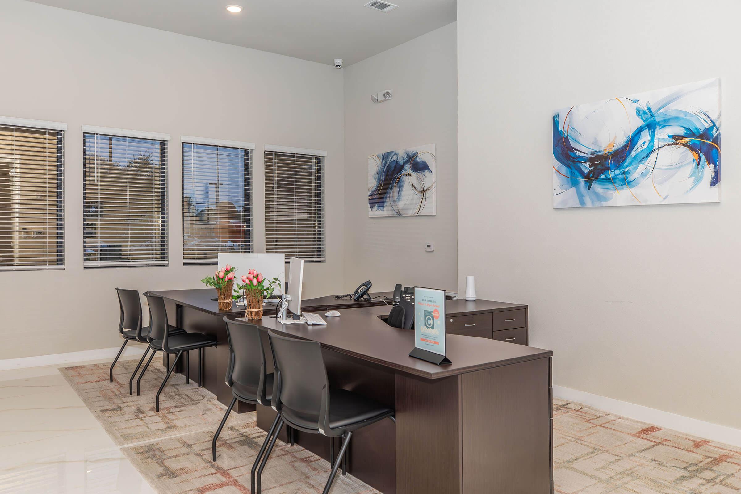 A modern office space featuring a long dark wooden desk with a phone and a tablet. There are four black chairs arranged around the desk. The walls are light-colored, and two abstract paintings in blue and white adorn one wall. Large windows with blinds allow natural light to fill the room. A beige area rug adds warmth to the space.