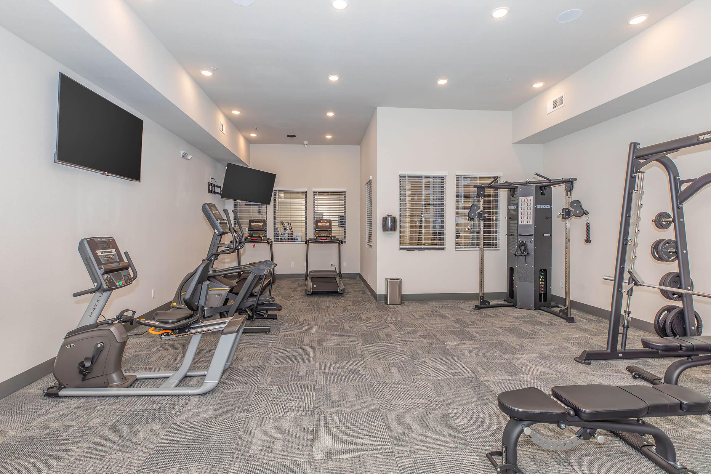 A well-lit gym area featuring exercise equipment including a treadmill, elliptical machine, multi-gym strength training equipment, and free weights. The walls are painted a light color, and there are two flat-screen TVs mounted for entertainment. The flooring is carpeted in a neutral shade.