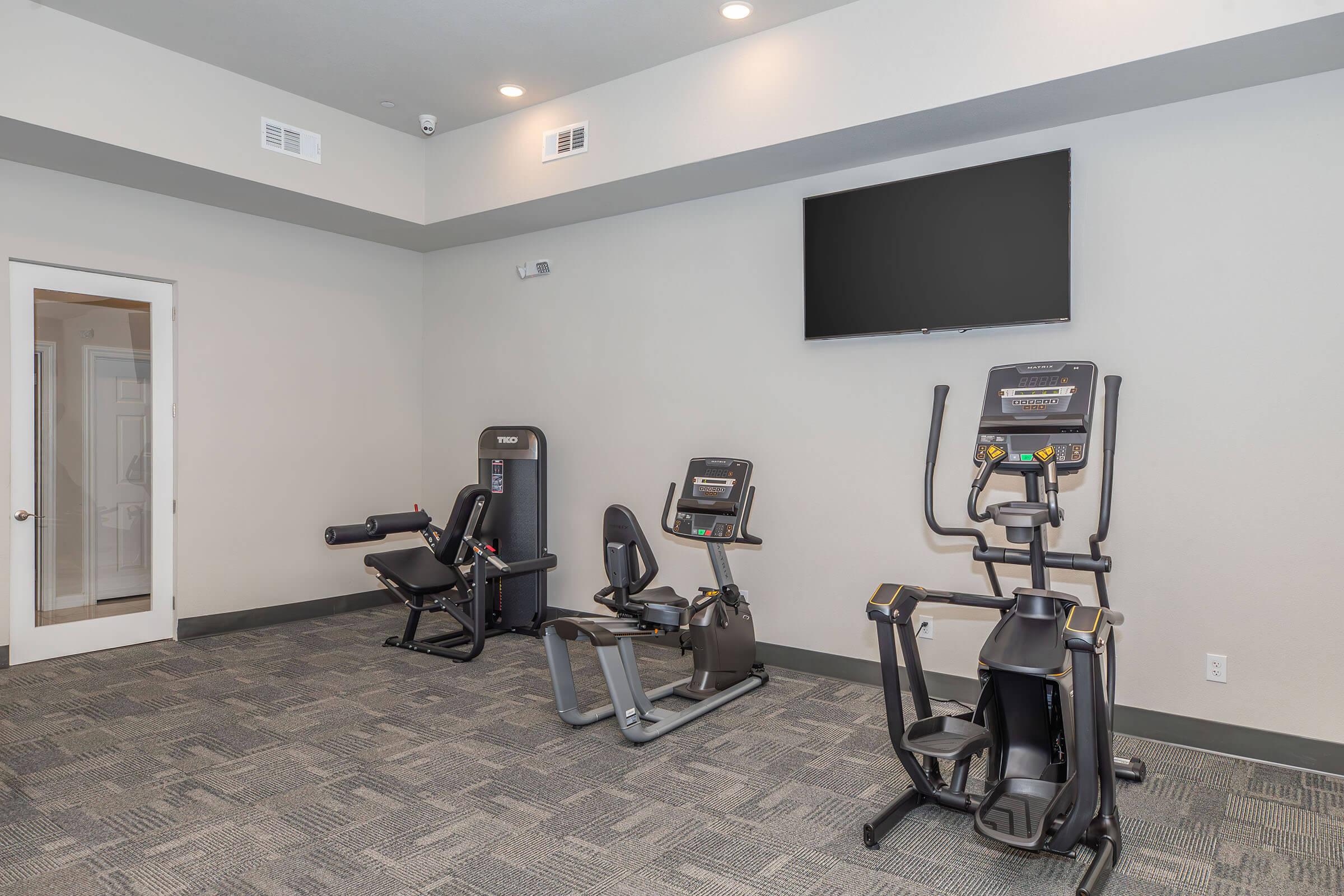 A modern gym space featuring exercise equipment including a recumbent bike, an elliptical machine, and a multi-station weight machine. The room is well-lit with overhead lights, has a large flat-screen TV mounted on the wall, and a door leading to another area. The flooring is carpeted with a neutral color scheme.