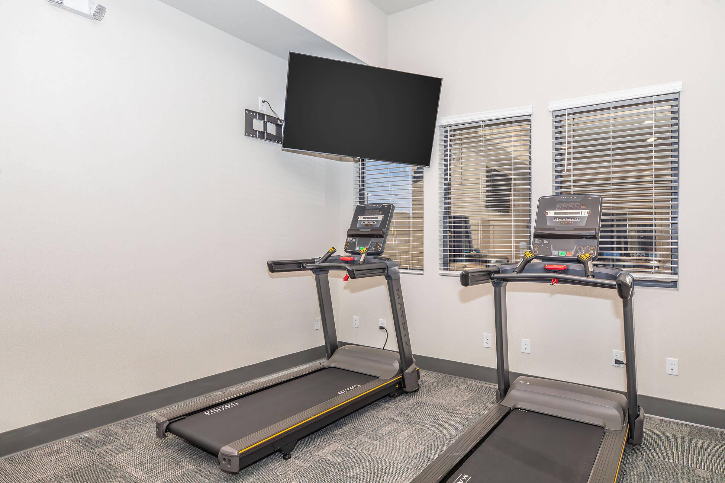 Two treadmills set up in a modern gym space with a large wall-mounted television above them. The room has large windows allowing natural light, and the flooring is a neutral gray carpet. The overall setting is clean and well-lit, designed for fitness activities.
