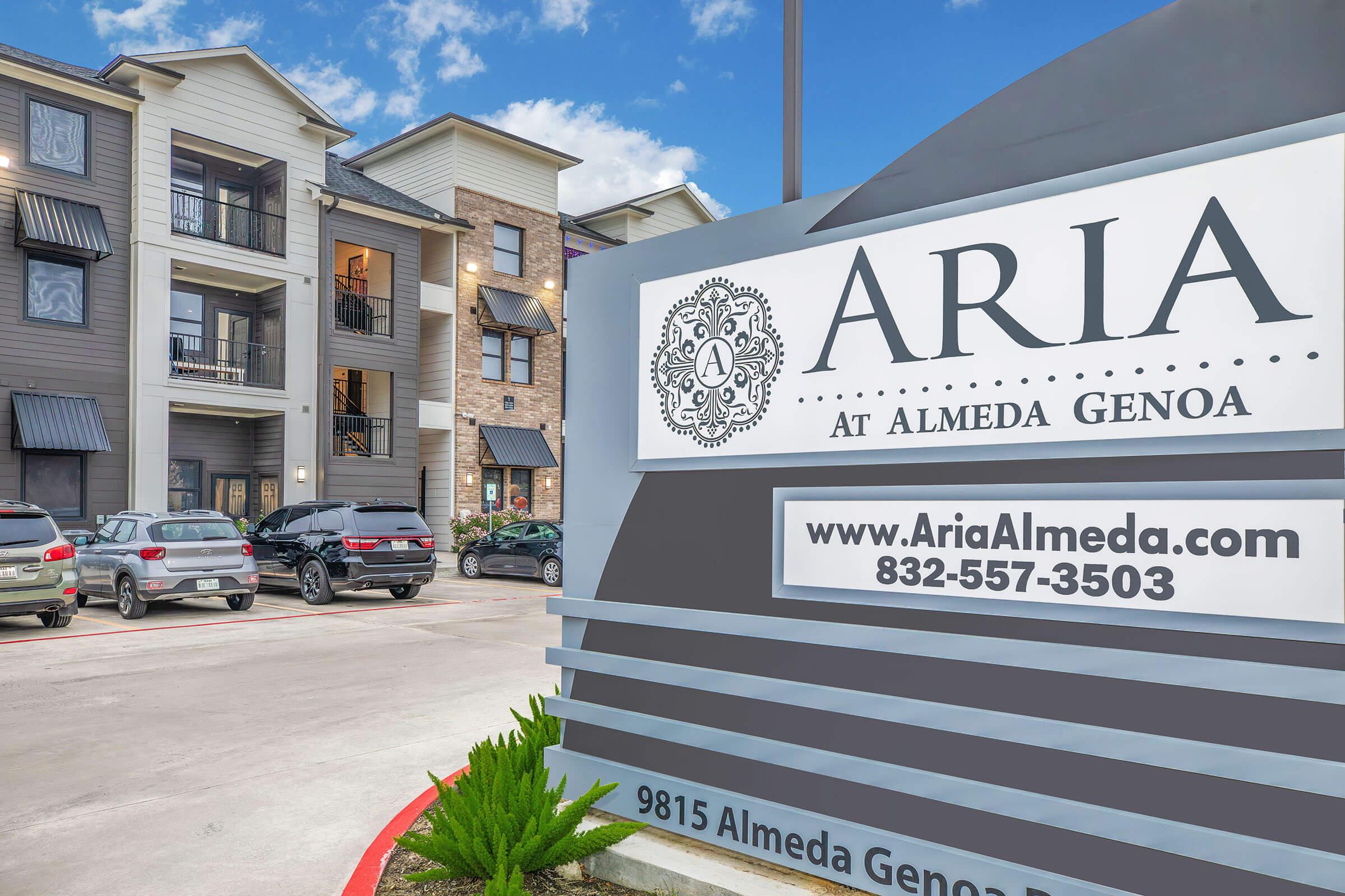 Exterior view of the Aria at Almeda Genoa apartment complex, featuring a modern design with multiple buildings. A prominent sign displays the name "Aria" alongside a website and contact number. Several parked cars are visible in the foreground, and the sky is partly cloudy.