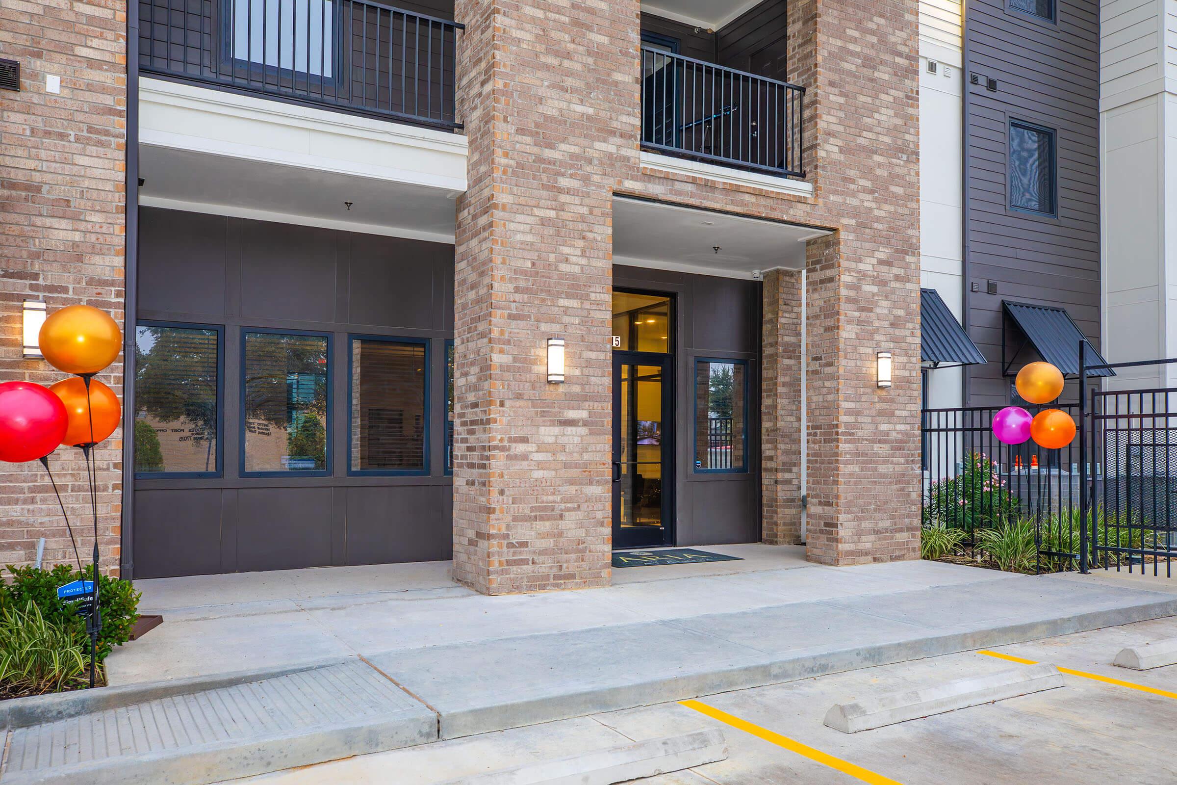 Exterior view of a modern apartment building entrance with a brick facade. The entrance features large windows, a double door, and decorative lighting. Colorful balloons in orange and pink are displayed on either side of the entrance, adding a festive touch. A paved area with parking spaces is visible in front.