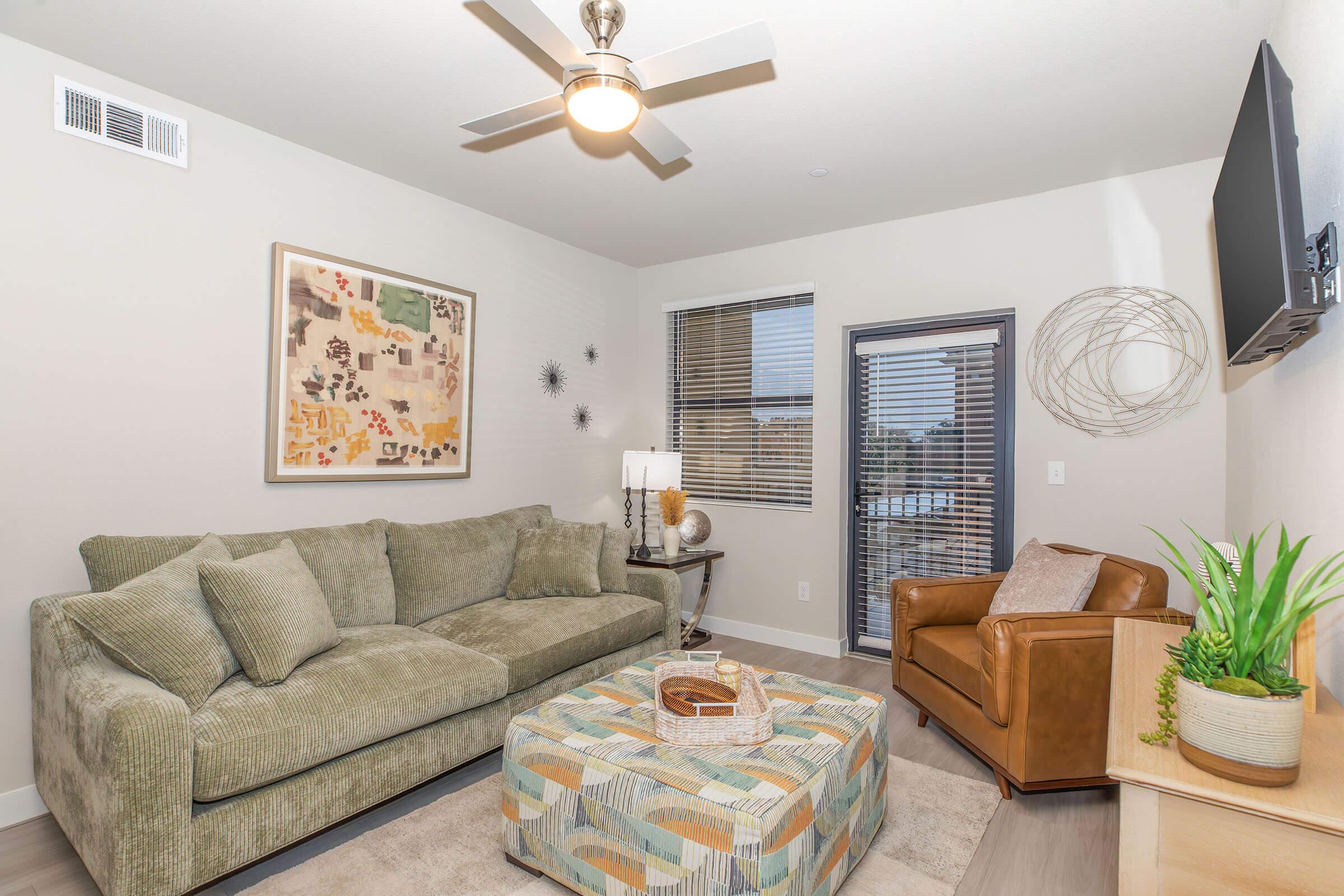 A modern living room featuring a green sectional sofa, a brown leather armchair, and a decorative coffee table with a patterned surface. A lamp illuminates the space, and large windows provide natural light. Wall decor includes an abstract artwork and a circular wall sculpture.