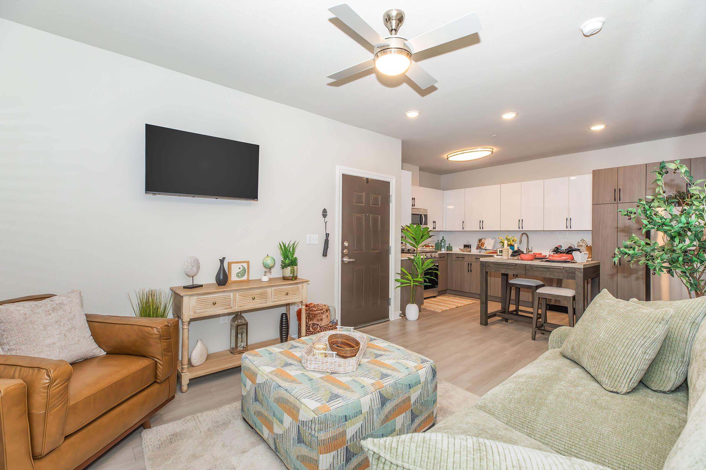 Cozy modern living room featuring a brown leather armchair, a textured green sofa, and a colorful patterned ottoman. A flat-screen TV is mounted on the wall. The open kitchen is visible in the background, showcasing white cabinets and a dining area with natural light and decorative plants.