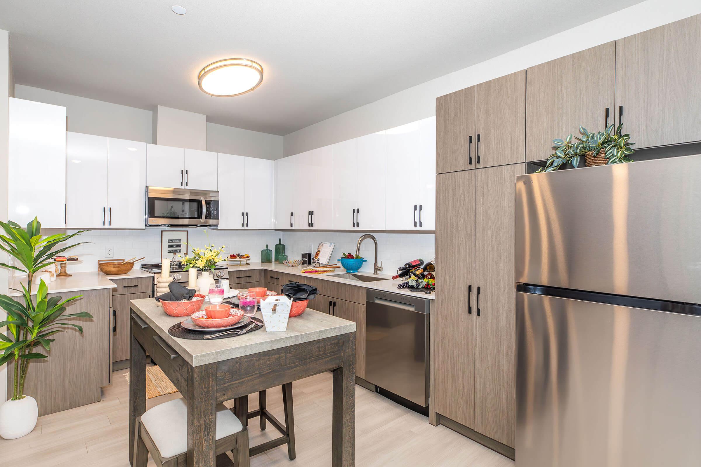 Modern kitchen featuring sleek cabinetry in light and dark wood tones. Includes stainless steel appliances, a stylish dining table set with colorful dishes, and decorative plants. Brightly lit with natural light, the space is organized and inviting, perfect for cooking and dining.