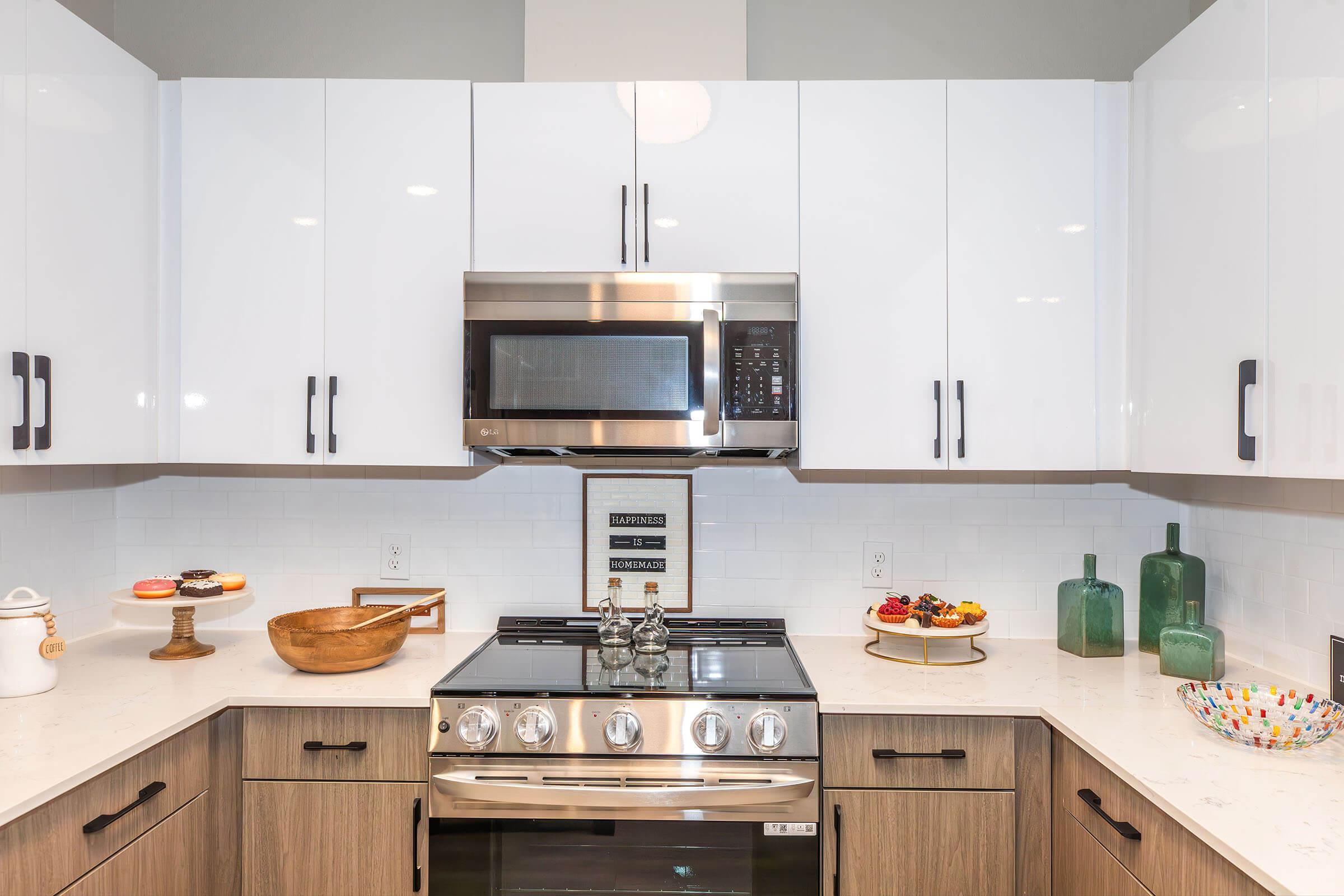 Modern kitchen featuring sleek white cabinets, a stainless steel microwave above a gas stove, and a countertop with a wooden bowl and decorative glass vases. A colorful dish holds assorted pastries, adding a touch of warmth to the contemporary design.
