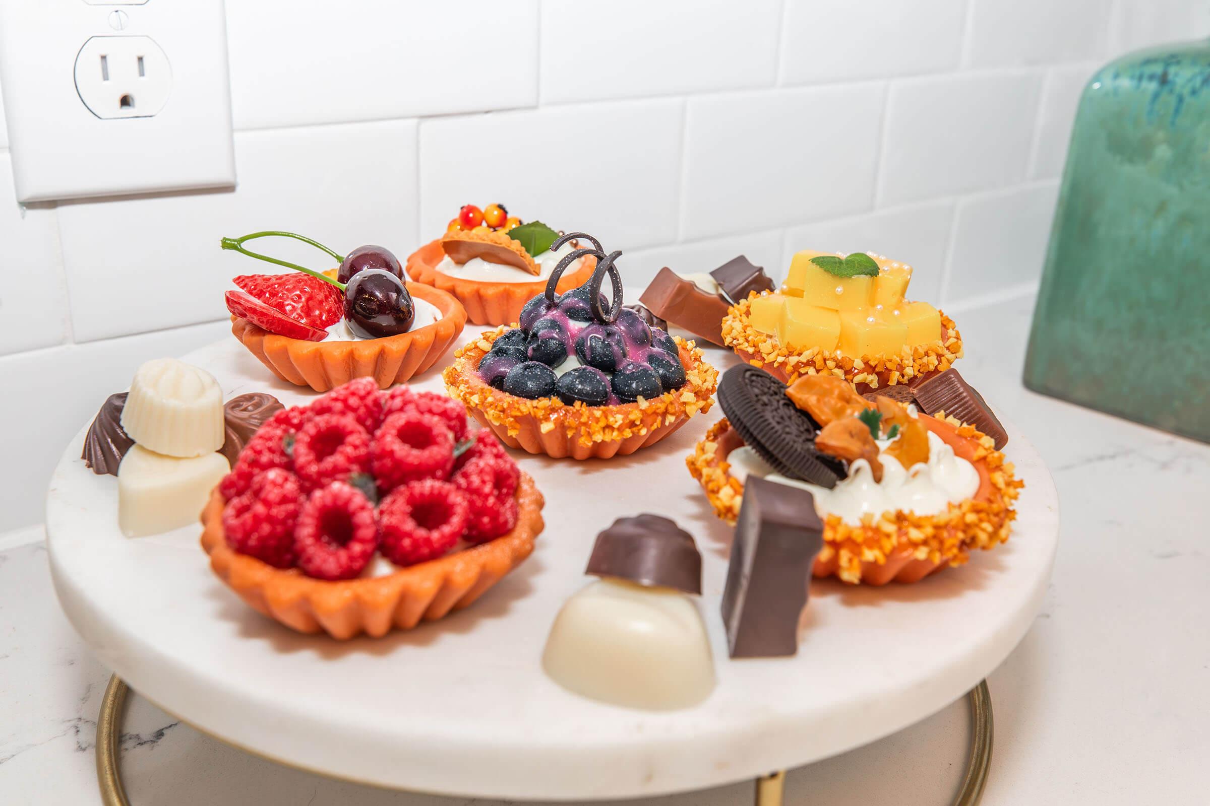 A platter displaying an assortment of colorful mini desserts, including chocolate-covered treats, fruit-topped tarts with raspberries, blueberries, strawberries, and cream, arranged on a white marble surface with a decorative golden stand.