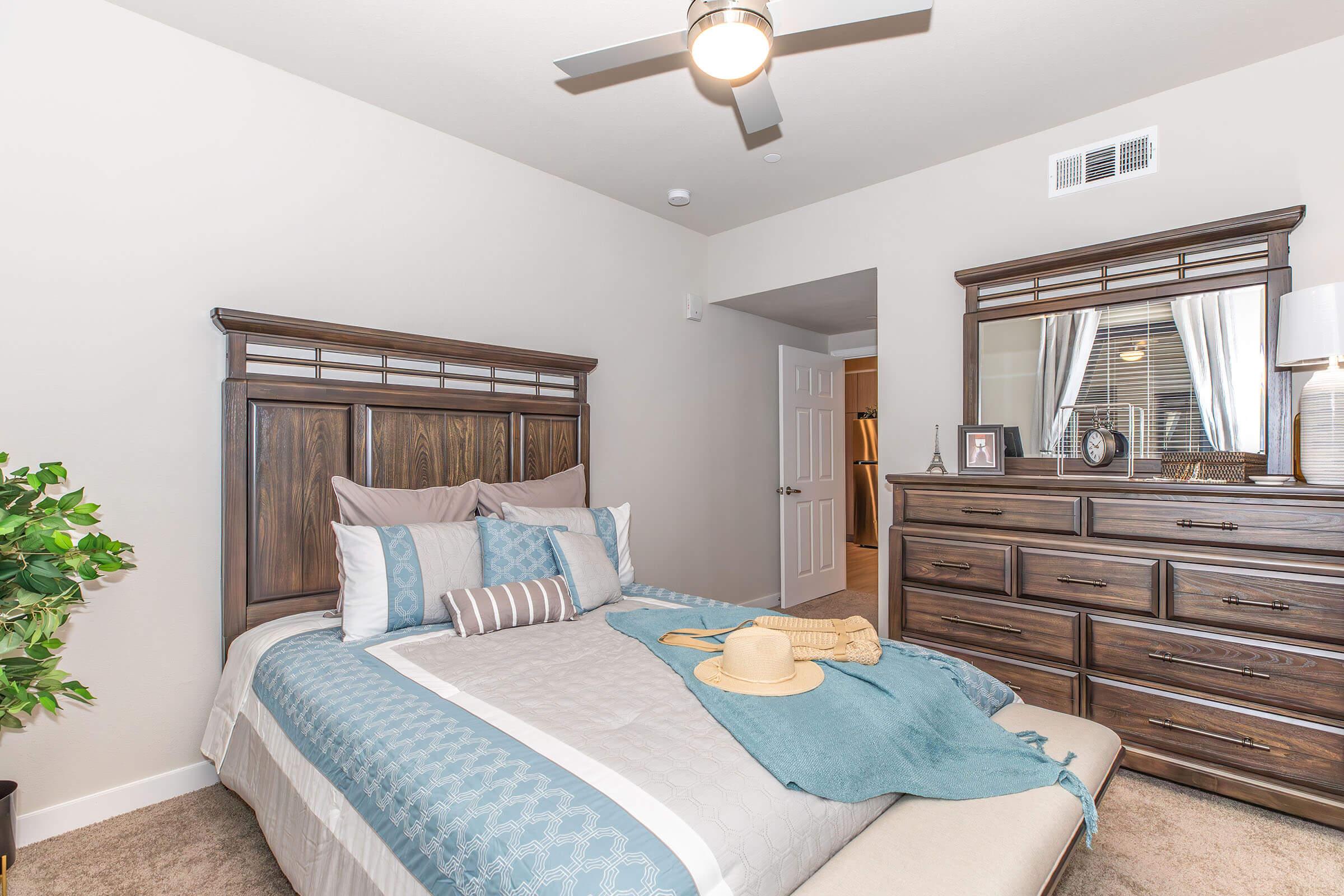A cozy bedroom featuring a wooden bed frame with decorative pillows, a turquoise throw blanket, and a straw hat placed on the bed. A dresser with a lamp and framed photo is positioned nearby, while a small indoor plant adds a touch of greenery. The walls are painted a soft color, enhancing the inviting atmosphere.