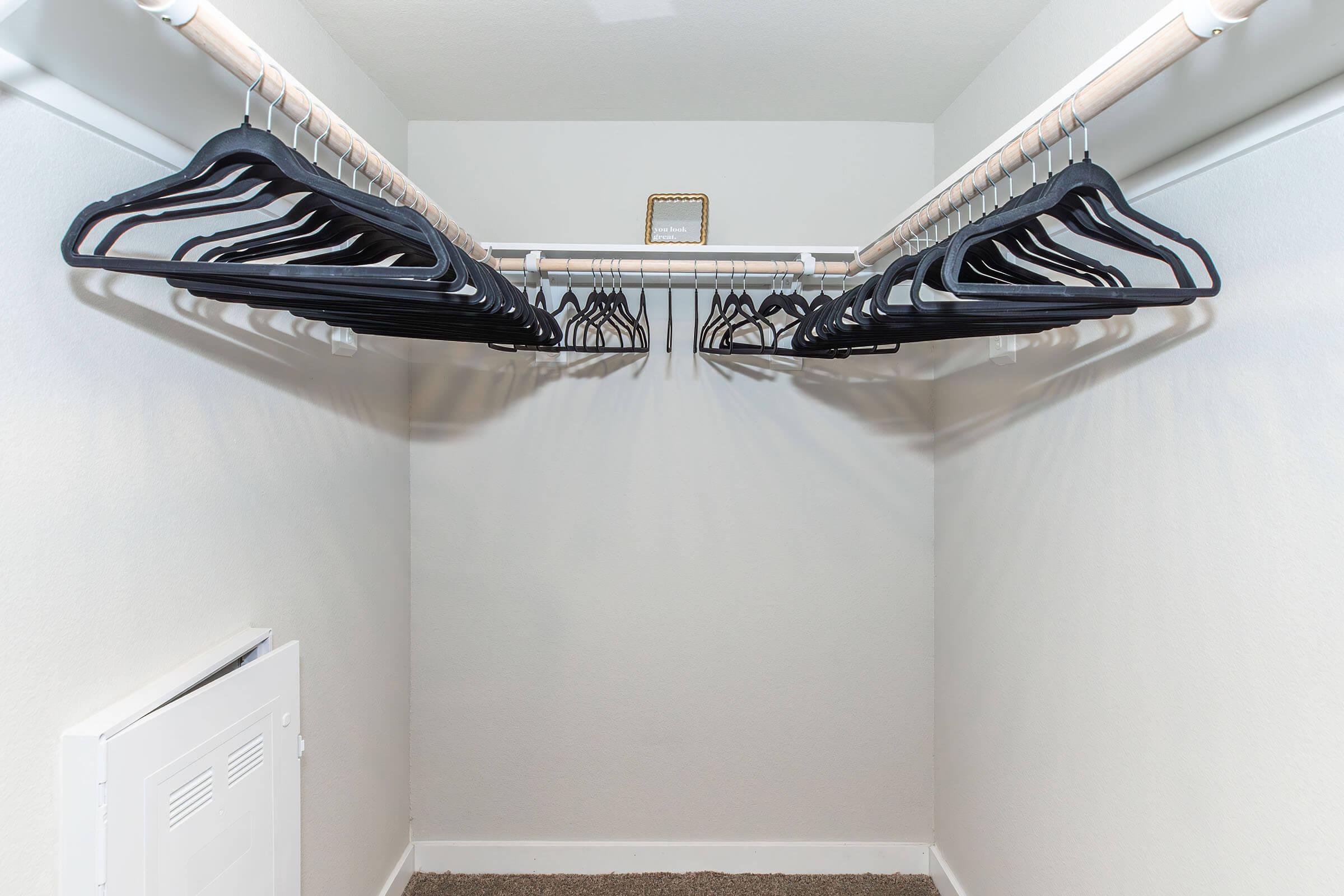 A minimalist closet interior featuring a white wall and a carpeted floor. There are multiple black hangers neatly arranged on a rod, with a small mirror placed on a shelf above. A white vent is visible on the left side of the image. The space is well-lit and orderly.
