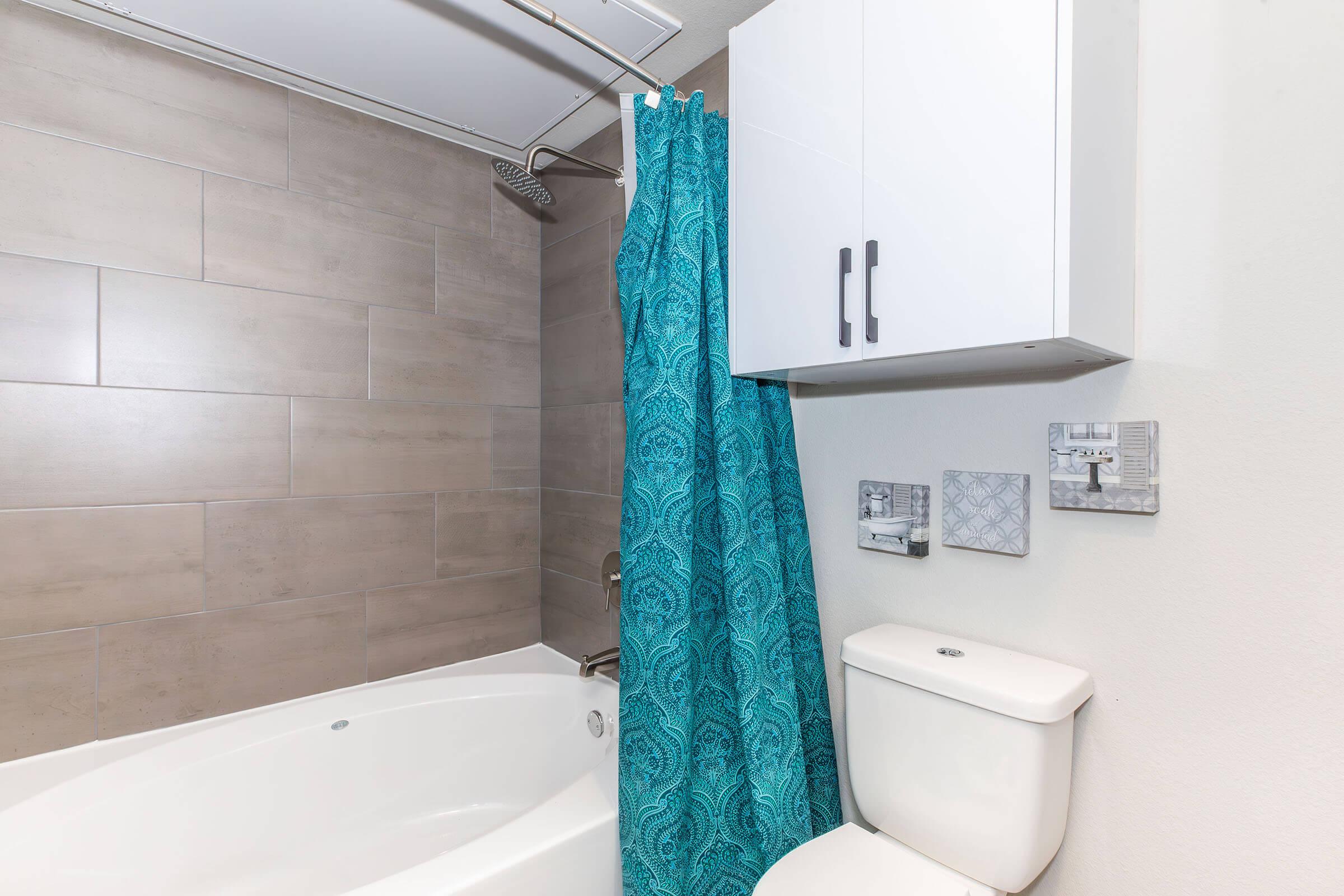 A modern bathroom featuring a curved bathtub with a light blue patterned shower curtain, a white toilet, and wall-mounted storage cabinets. The walls are tiled with light gray porcelain tiles, and the overall decor is clean and contemporary.