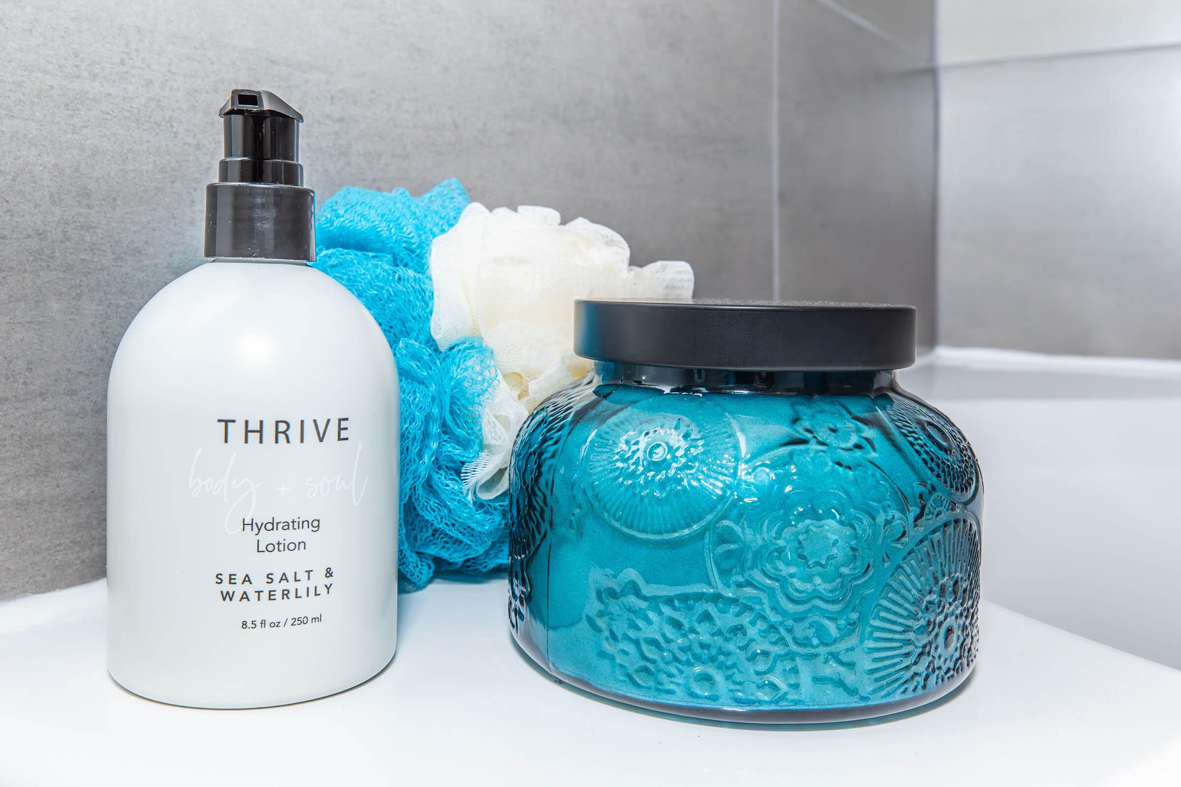 A white bottle of Thrive Hydrating Lotion with a black pump, labeled "Sea Salt & Waterlily," next to a decorative blue jar with an embossed floral pattern. A light blue shower loofah is resting in the background, on a gray tiled surface. The scene is set in a bright bathroom.