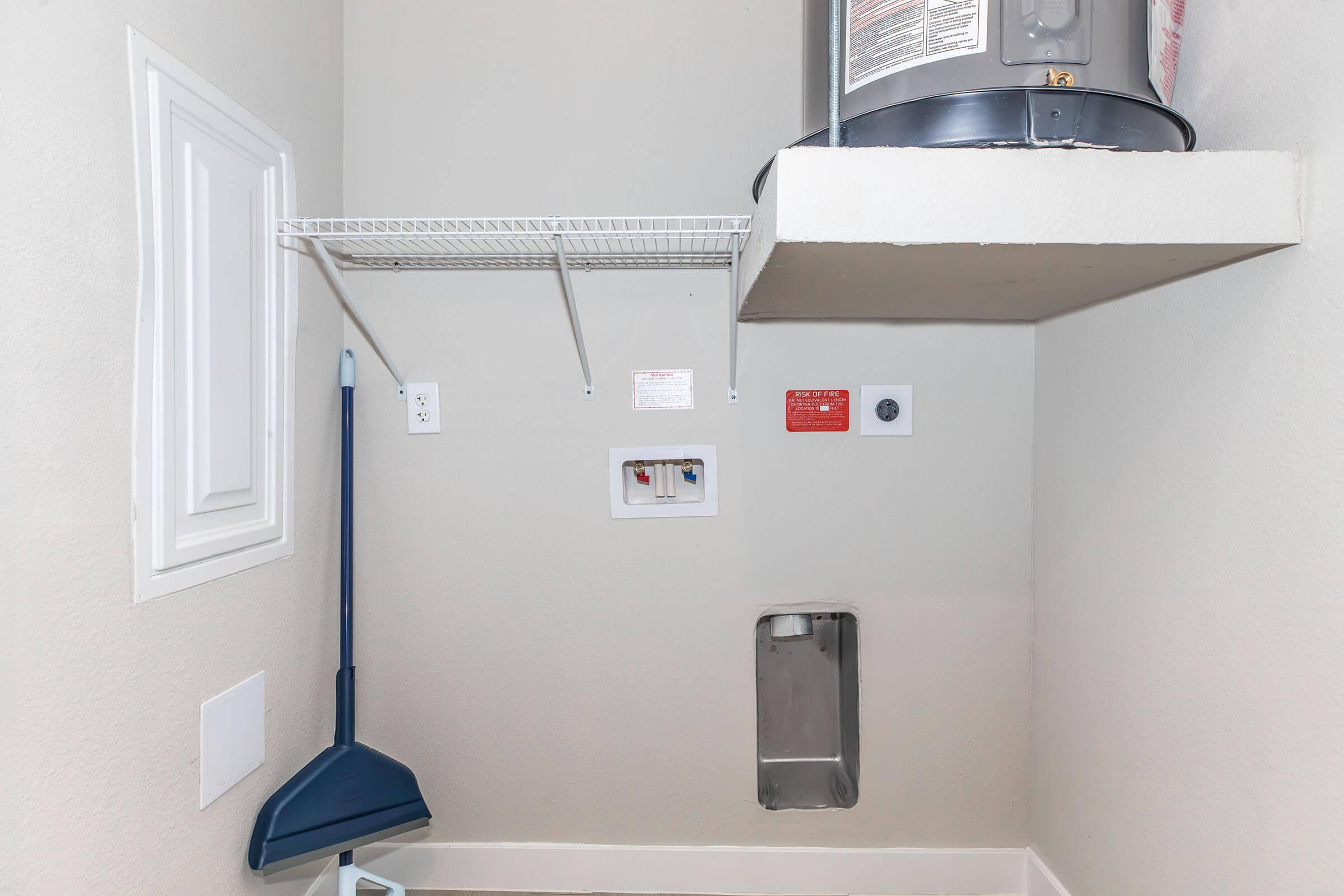 A clean and minimal utility room featuring a wall-mounted water heater, a shelving unit, a broom, and various electrical outlets. The walls are painted in a light color, and there is a small access panel on the floor.