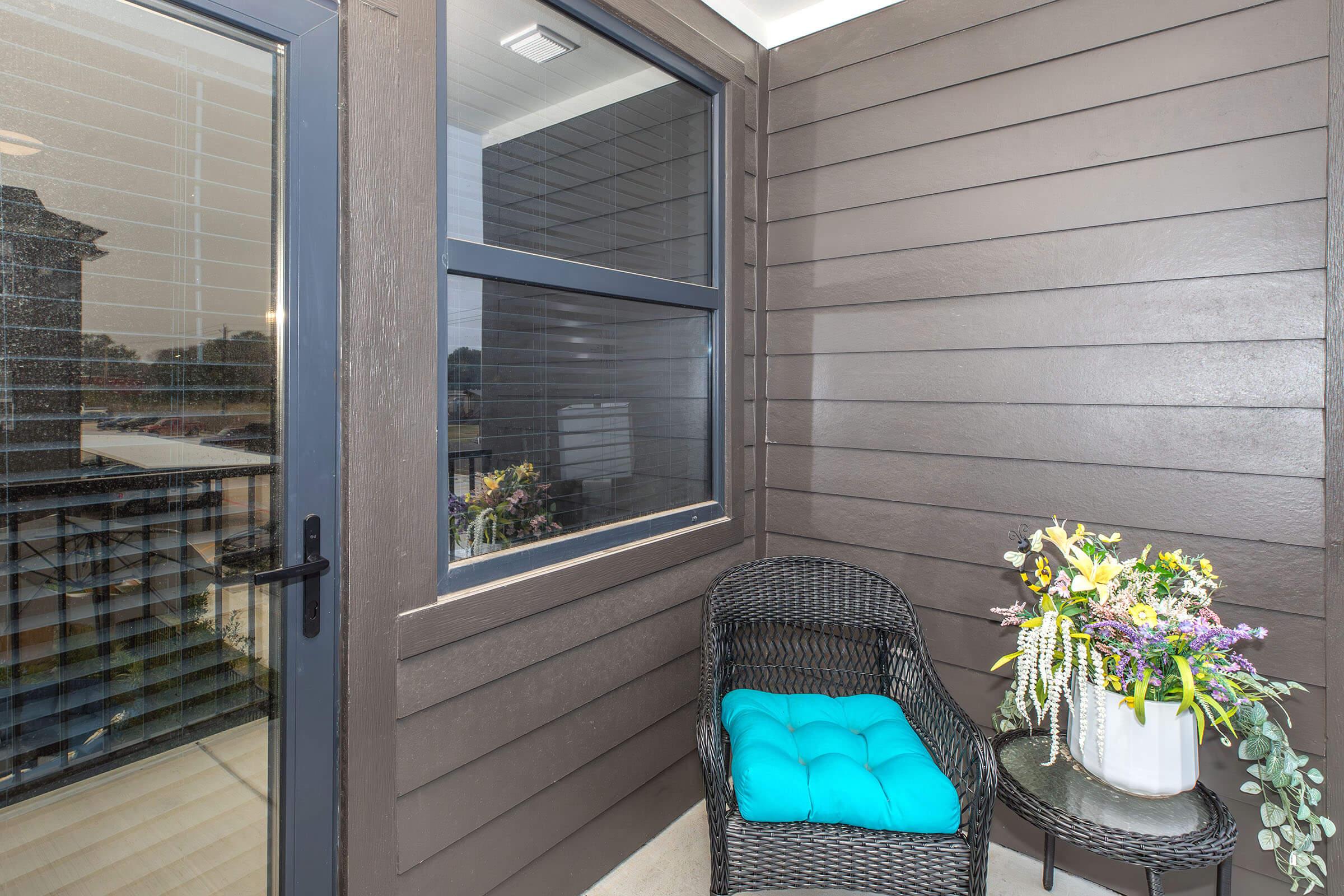 A small balcony featuring a wicker chair with a blue cushion and a round table adorned with a bouquet of flowers. The walls are painted a rich brown, and a sliding glass door provides access to the area. Natural light filters in, creating a cozy atmosphere.