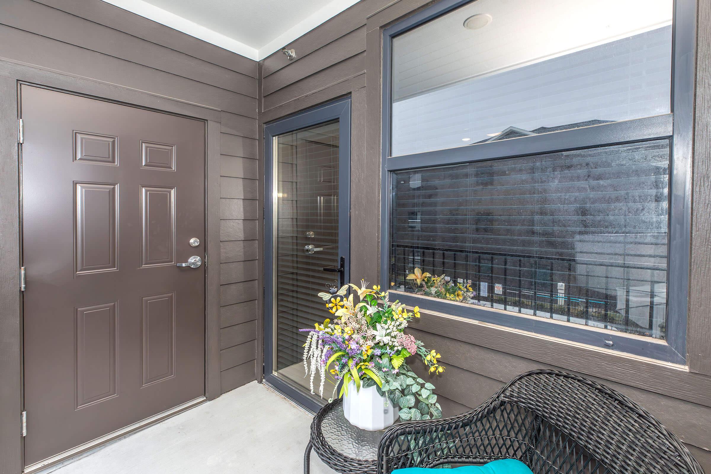 A cozy entrance area with a dark brown door and a glass-paneled door leading into the home. A small table with a bouquet of flowers sits on the right, alongside a wicker chair. The walls are clad in dark wood siding, and a glimpse of the outdoors is visible through the window.