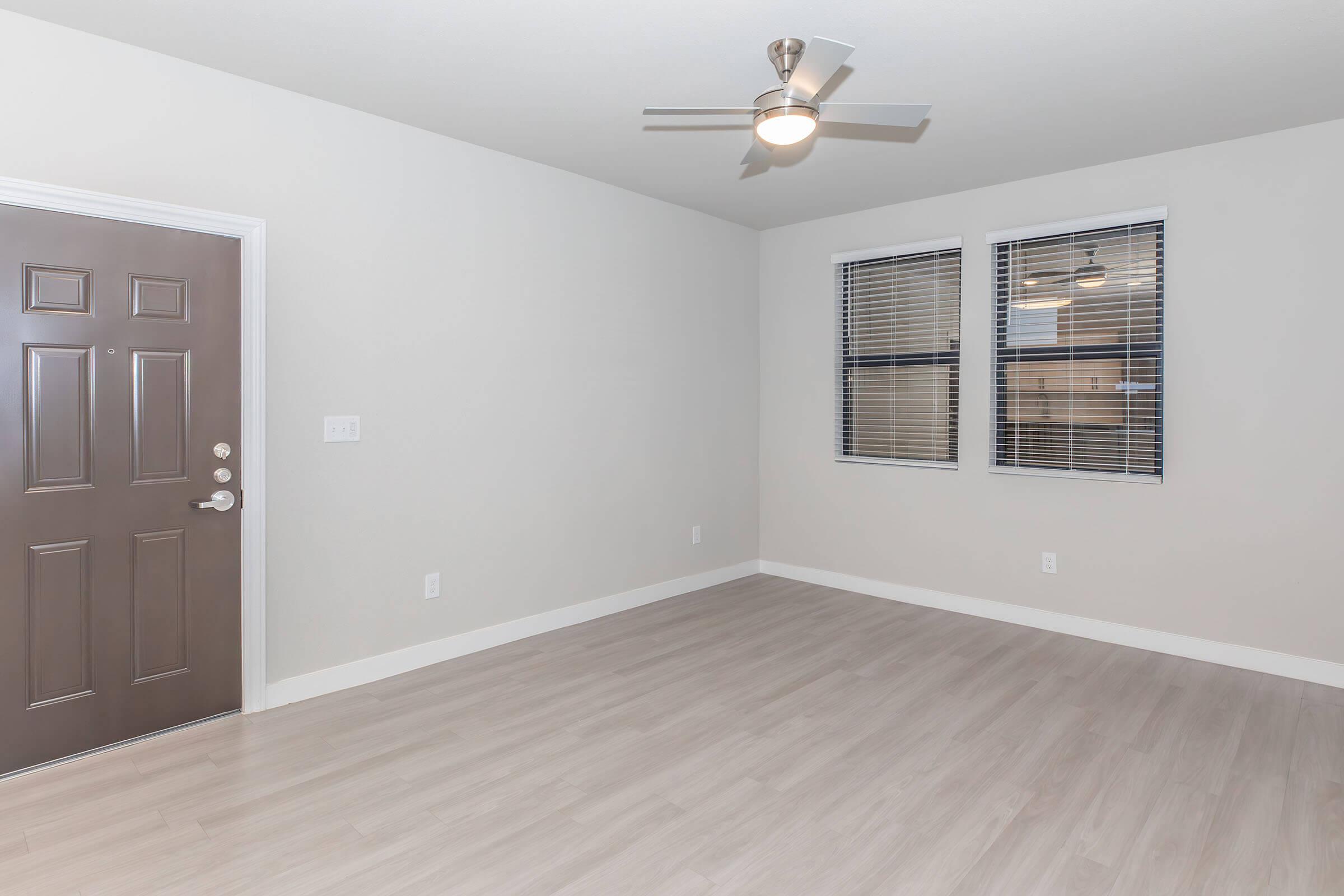 A spacious, empty living room featuring a brown front door, two large windows with blinds, and a ceiling fan. The floor is light-colored laminate, and the walls are painted a neutral beige. The room is well-lit with natural light coming in through the windows.