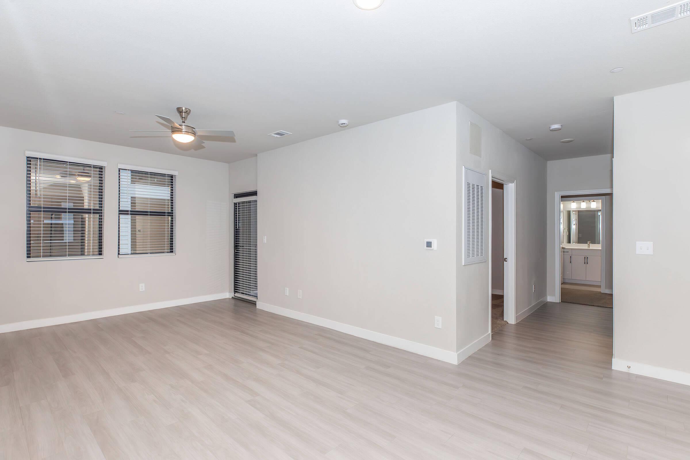 Bright and airy interior of a living room with light wood floors, light gray walls, and large windows with blinds. A ceiling fan is mounted on the ceiling. A doorway leads to a bathroom area in the background, and there is a closet space visible to the left. Overall, the space appears modern and welcoming.