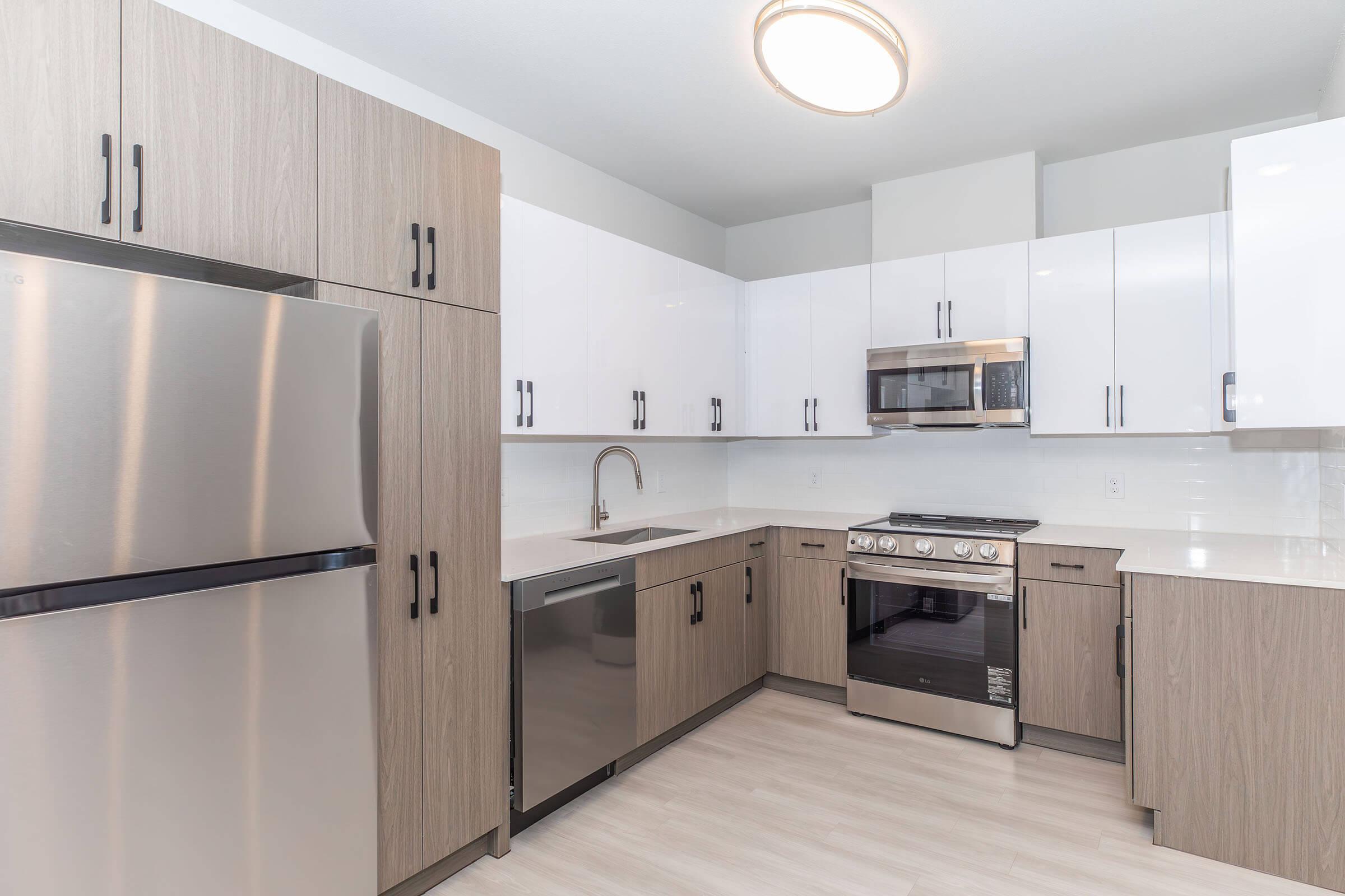 Modern kitchen featuring stainless steel appliances, including a refrigerator, microwave, and oven. The cabinetry has a mix of light and dark wood finishes. The countertop is white, and there is a sleek sink with a stylish faucet. Ample storage space is provided with a clean and contemporary layout.