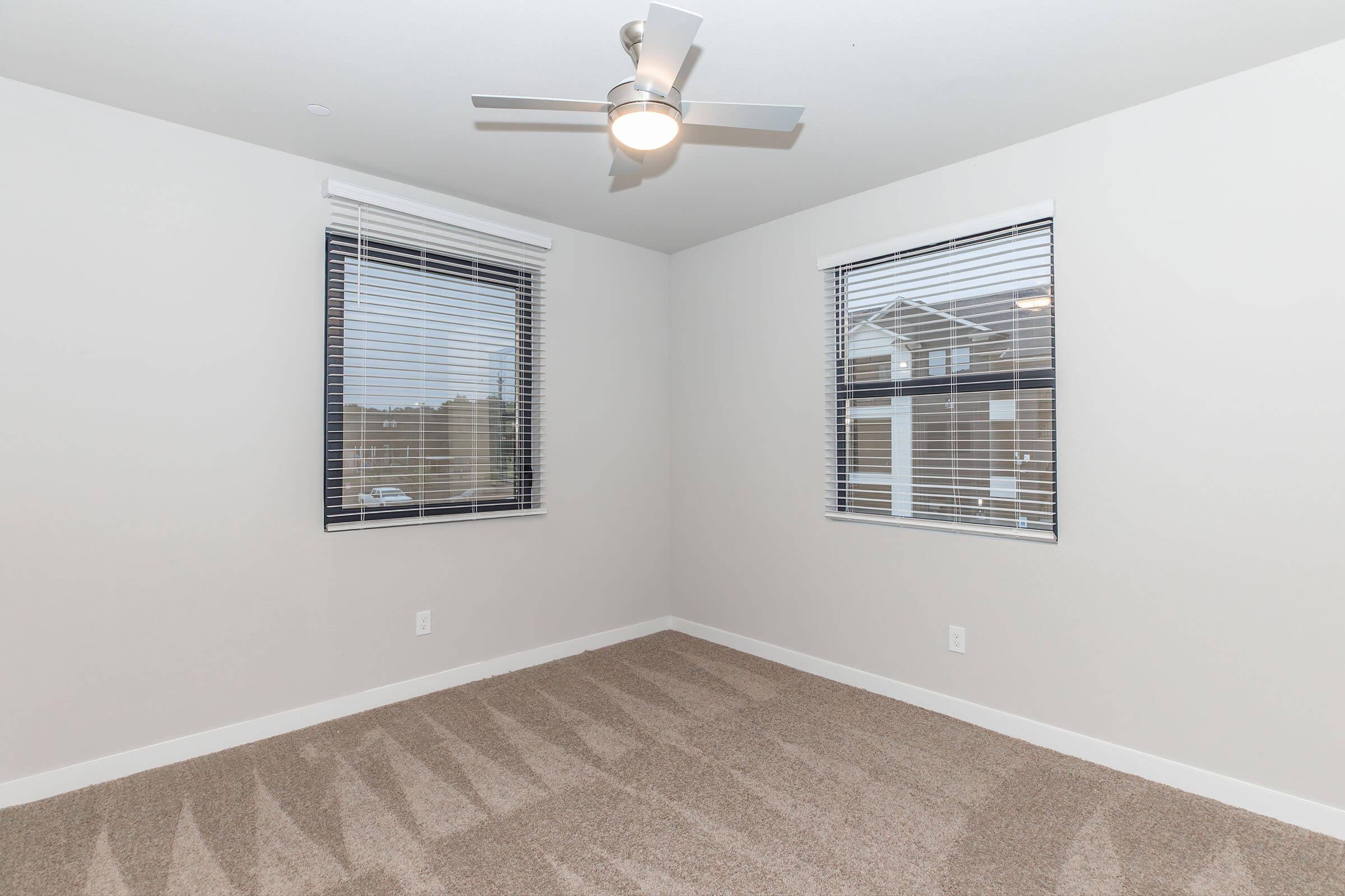 Empty room with light gray walls and a tan carpet. Two windows with white blinds provide natural light, and a ceiling fan with a light fixture is mounted in the center of the ceiling. The space is clean and uncluttered, ideal for various uses such as a bedroom or office.