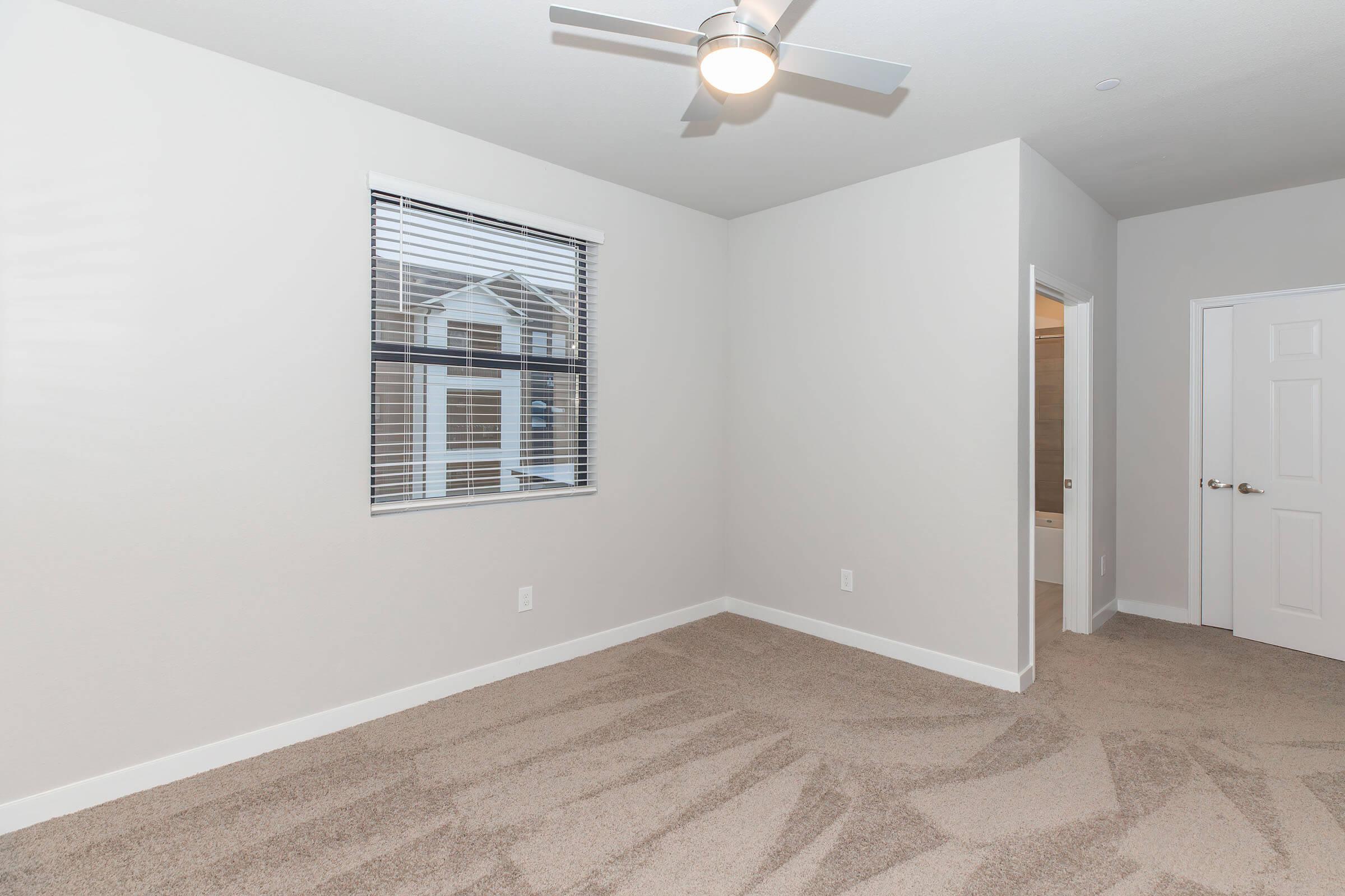 A bright, empty room with light gray walls and beige carpet. There is a window with blinds allowing natural light, a ceiling fan, and a doorway leading to a bathroom. The overall atmosphere is clean and modern, suitable for various interior design styles.