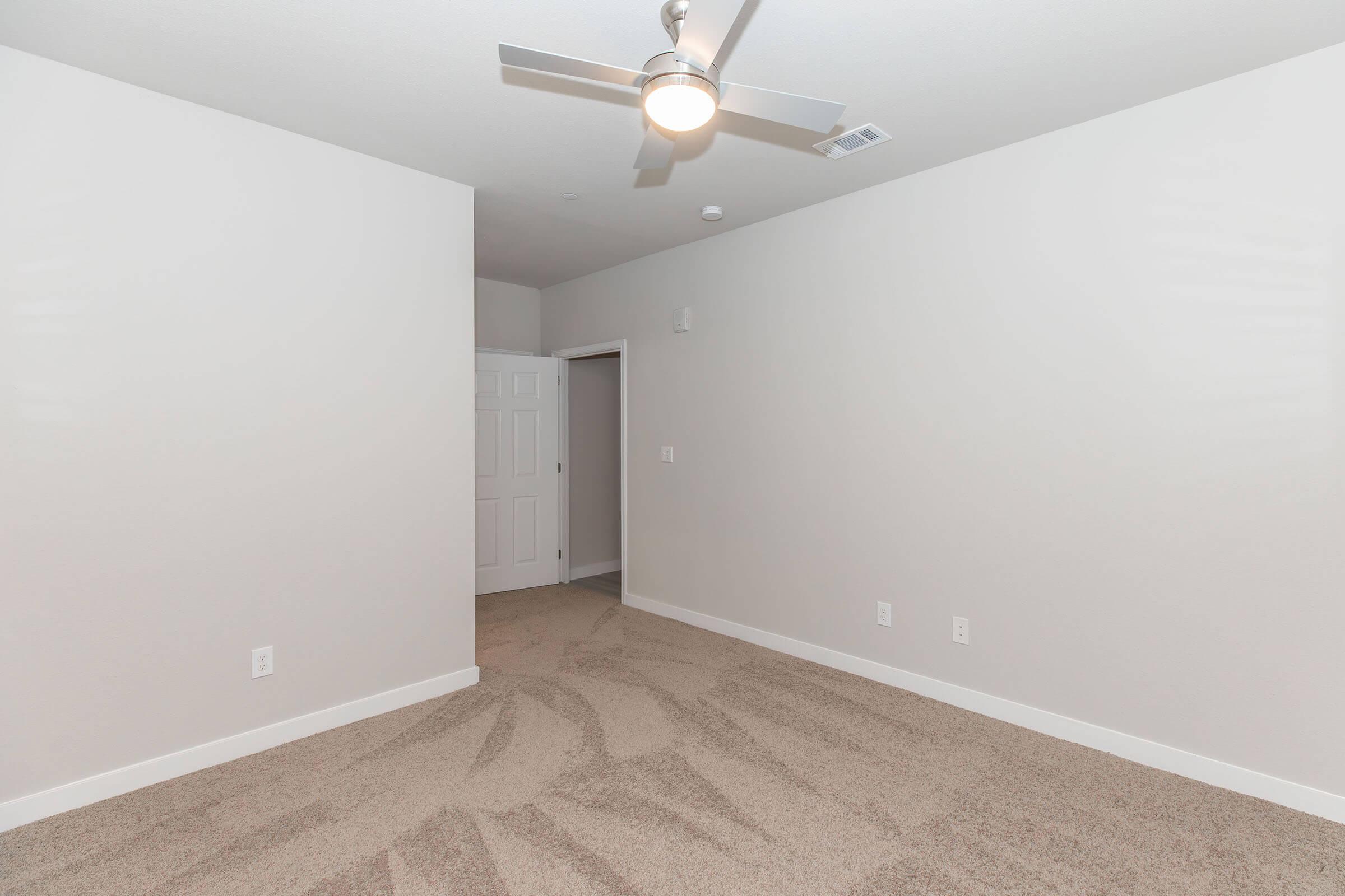 A spacious, empty room featuring light gray walls and carpeted flooring. A modern ceiling fan is mounted on the ceiling. The room has an open layout with a doorway leading to another area, creating a bright and inviting atmosphere.