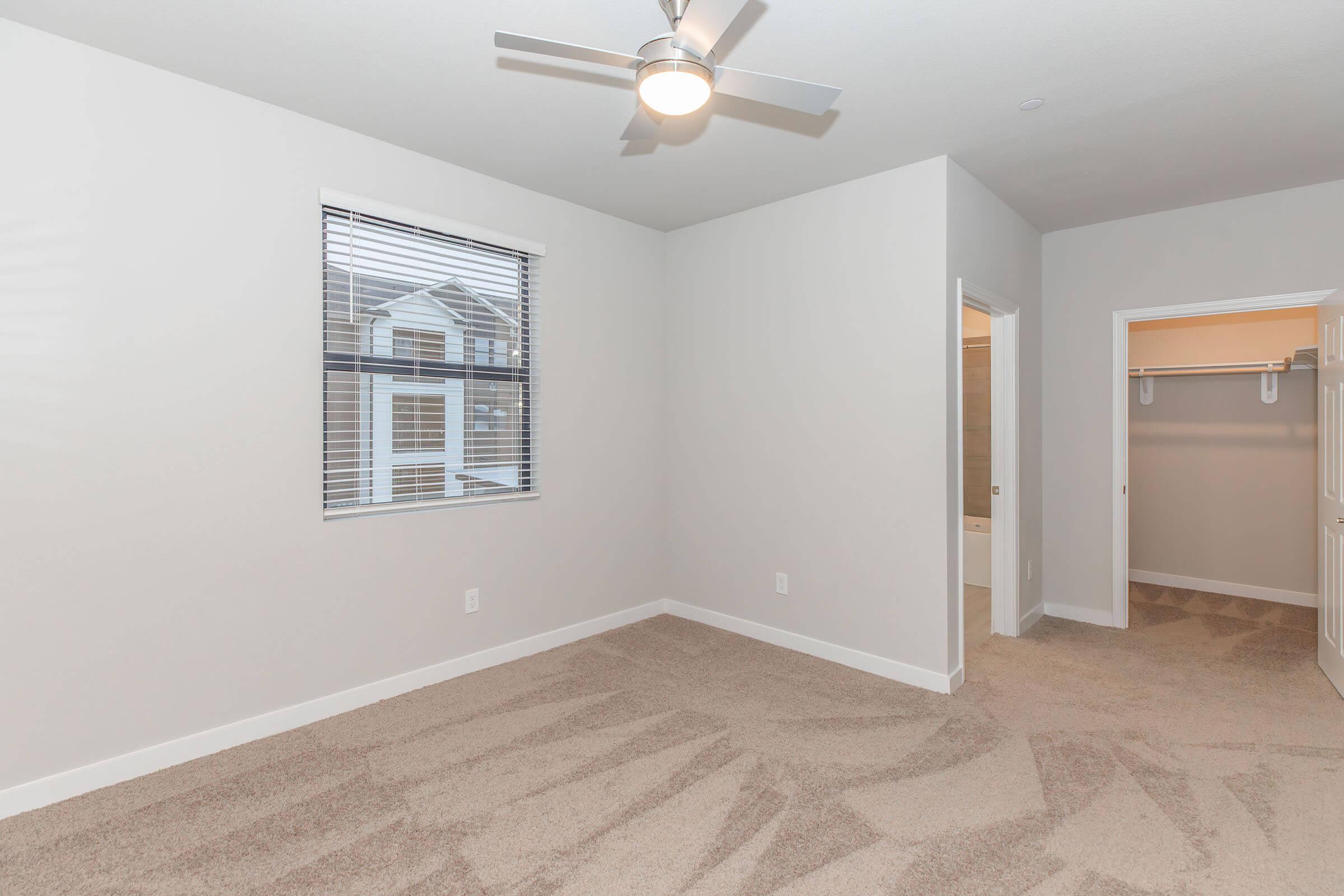 A bright, empty bedroom with light gray walls and beige carpet. It features a ceiling fan and a window with blinds allowing natural light. An open doorway leads to a bathroom, while another door reveals a closet space. The room has a modern, clean look with minimal furnishings.
