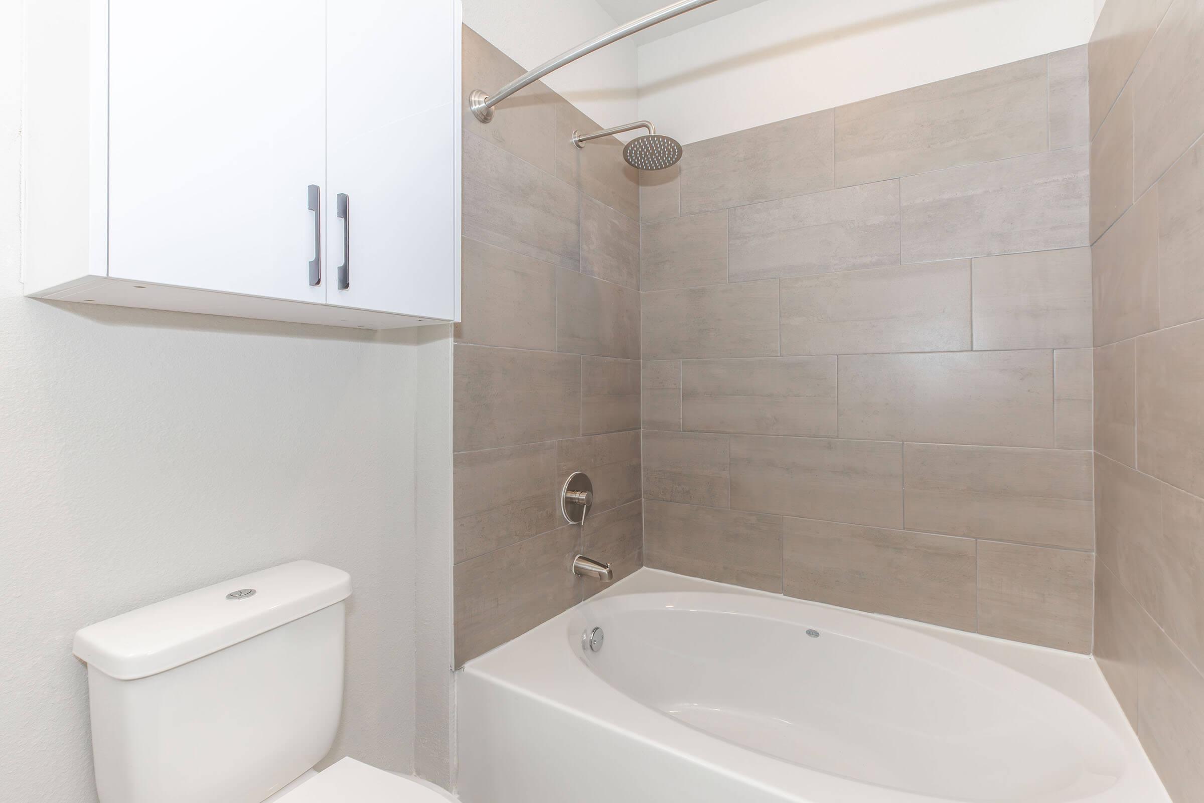A clean bathroom featuring a bathtub with a showerhead, light gray tiled walls, and a white toilet. A modern storage cabinet is mounted above the bathtub, enhancing the overall aesthetic of the space. The design emphasizes simplicity and functionality.