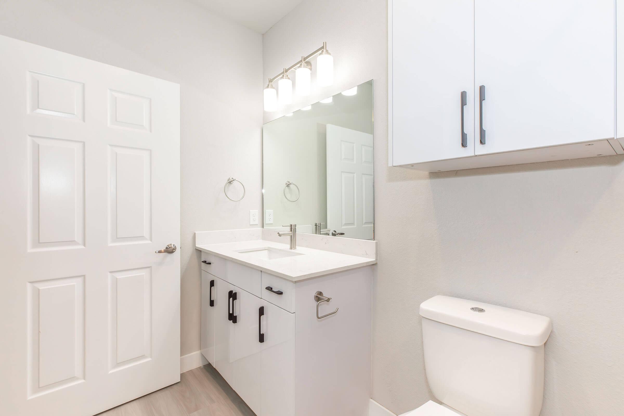 Modern bathroom featuring a white vanity with a sink and silver fixtures, a large mirror above, light fixtures, and a white toilet. The walls are light gray, and there's a closed white door visible. The overall aesthetic is clean and contemporary.