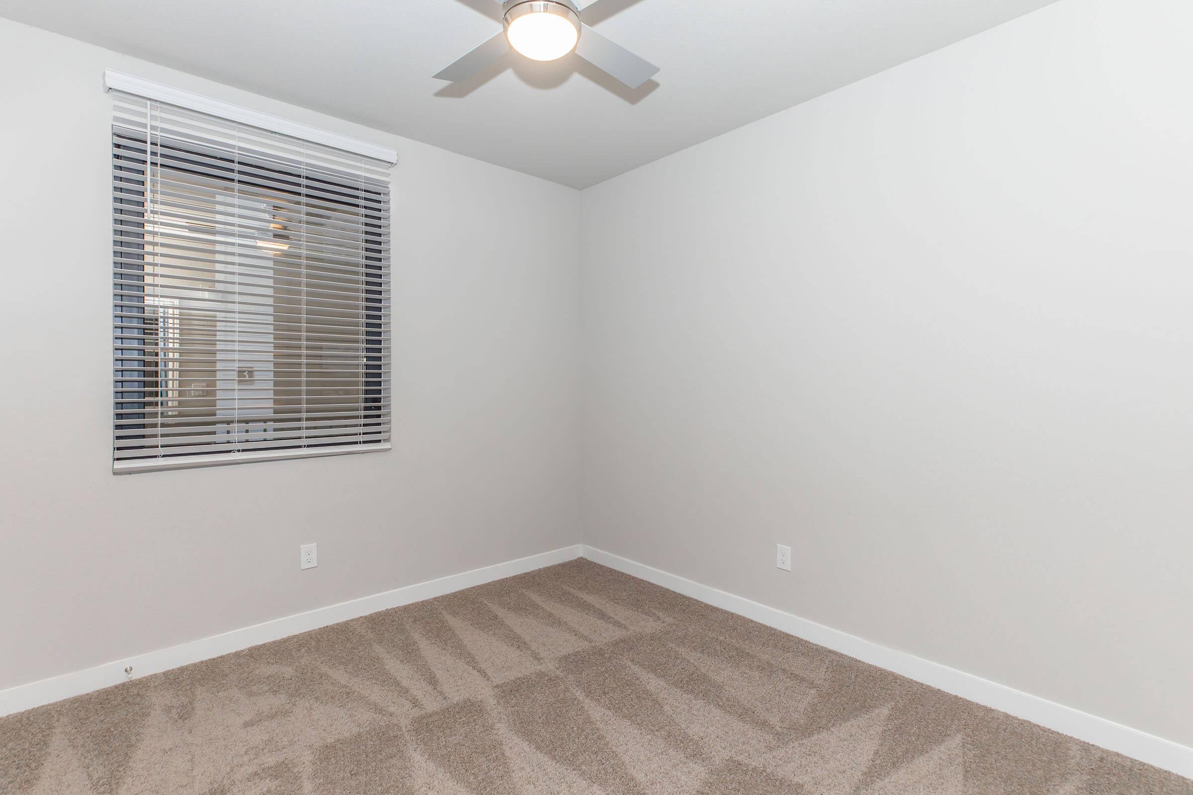 An empty room with light-colored walls and a ceiling fan. There is a window with white blinds on one wall, and the floor is covered with beige carpet. The room is well-lit and features a modern, minimalist aesthetic.