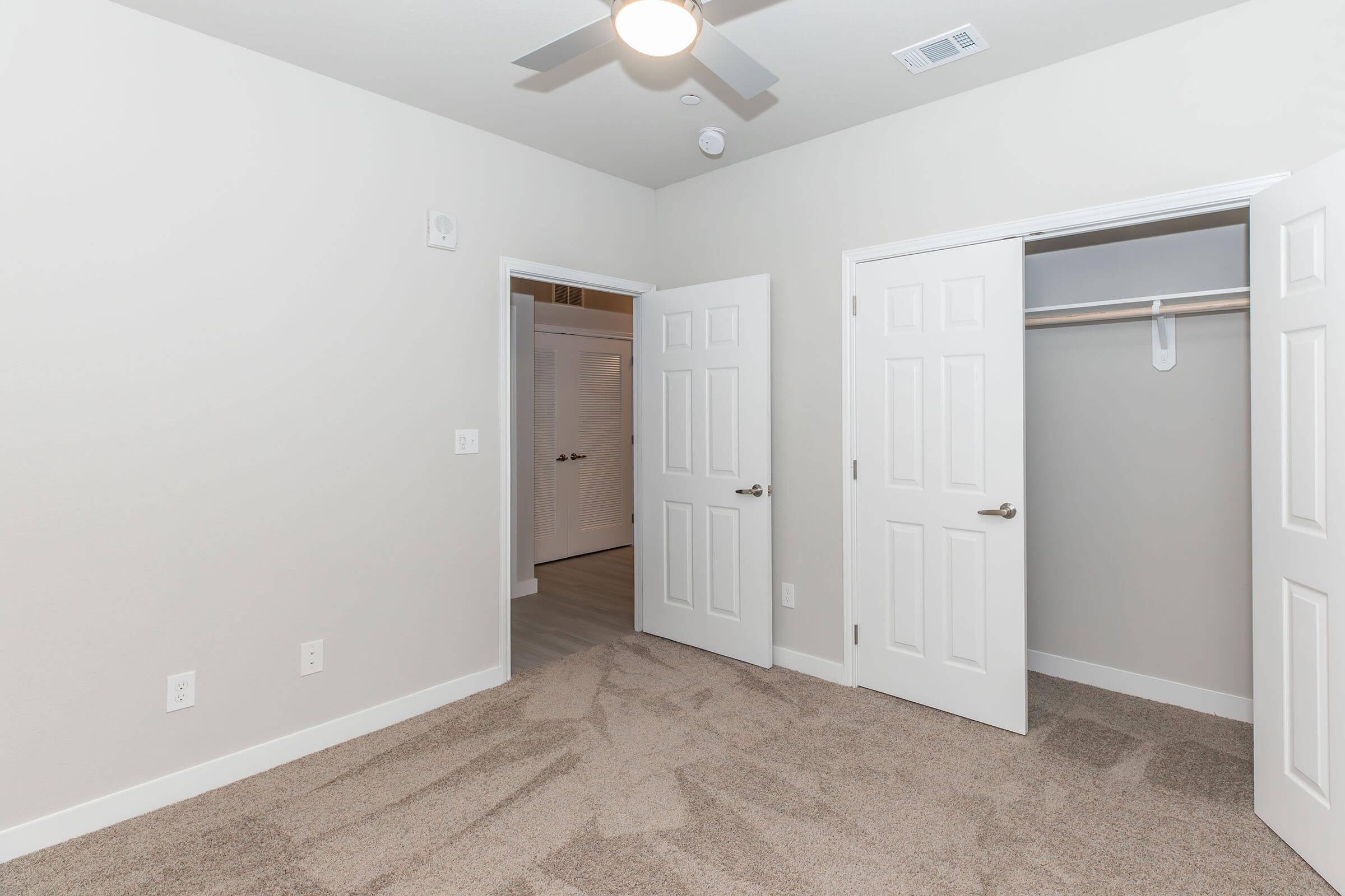 Interior view of a small, empty bedroom featuring light gray walls, plush carpet, and two white doors—one leading to a closet and the other to another room. A ceiling fan is visible above, and natural light streams from the doorway, creating a bright and open atmosphere.