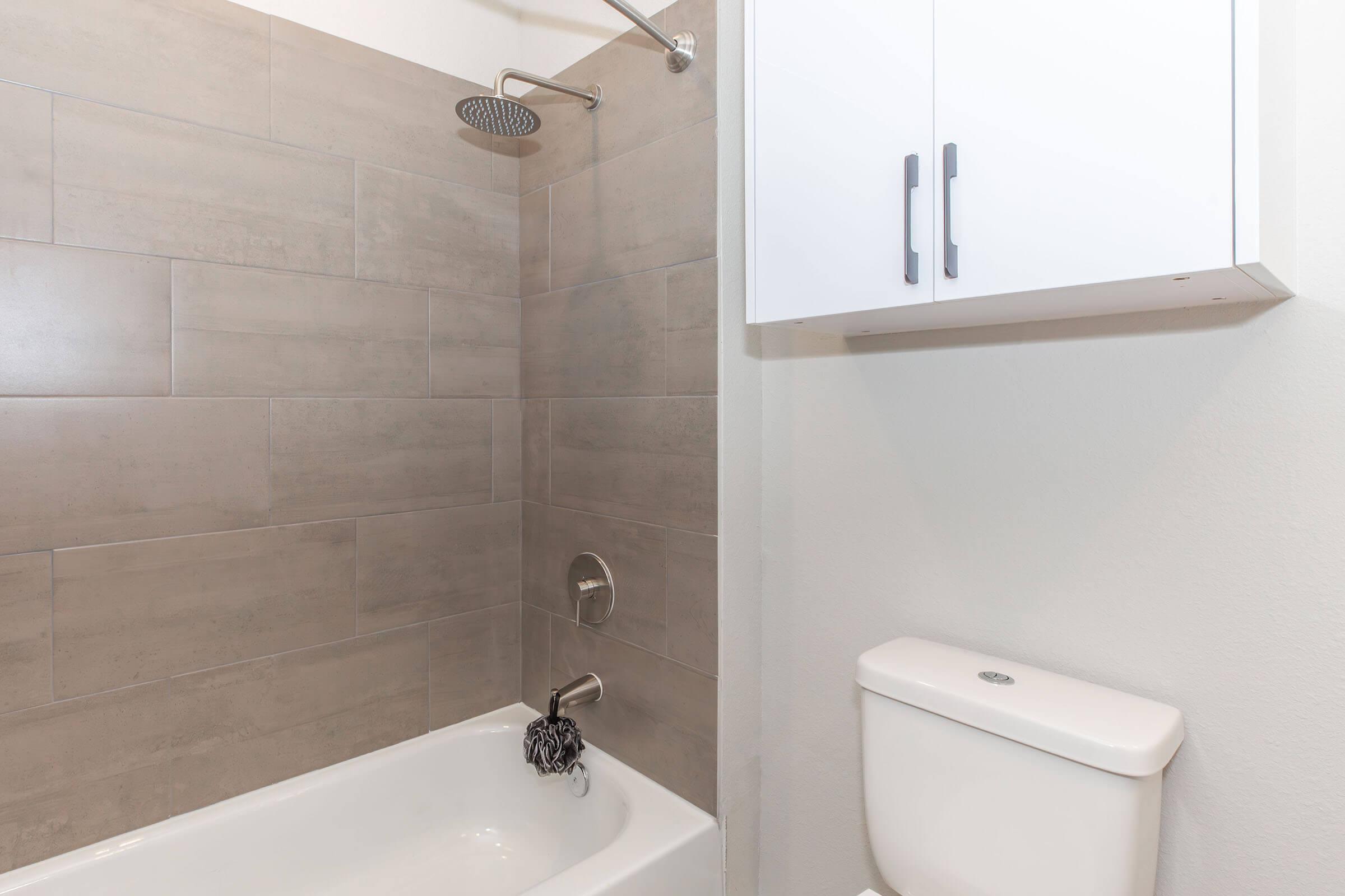 A modern bathroom featuring a shower with a sleek rain showerhead and tiled walls, a white bathtub, a toilet, and a wall-mounted cabinet with two doors for storage. The overall design is clean and minimalist, with neutral tones.