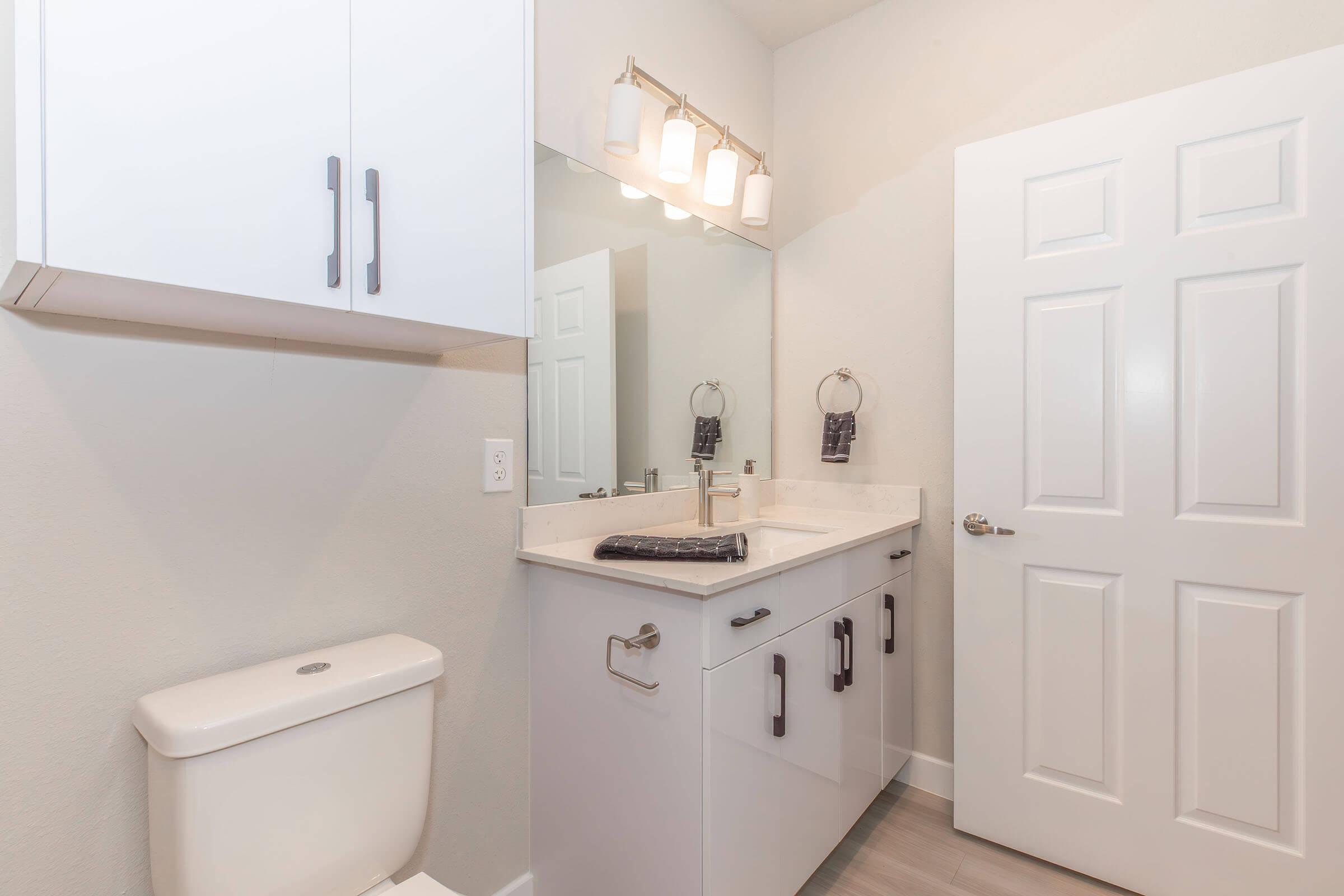 A modern bathroom featuring a white vanity with sleek black handles, a large mirror above a countertop, and a stylish light fixture. The space includes a white toilet and gray walls, creating a clean and contemporary look. A towel is hanging on a ring beside the sink.
