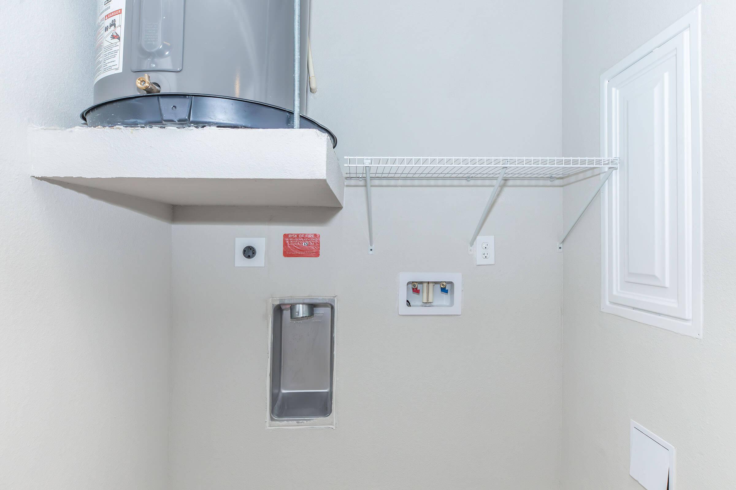 A small utility room featuring a water heater mounted on the wall, a wire shelving unit, and various electrical outlets and fixtures on the beige wall. There is a recessed panel and a small opening possibly for plumbing access. The overall space is well-lit and uncluttered.