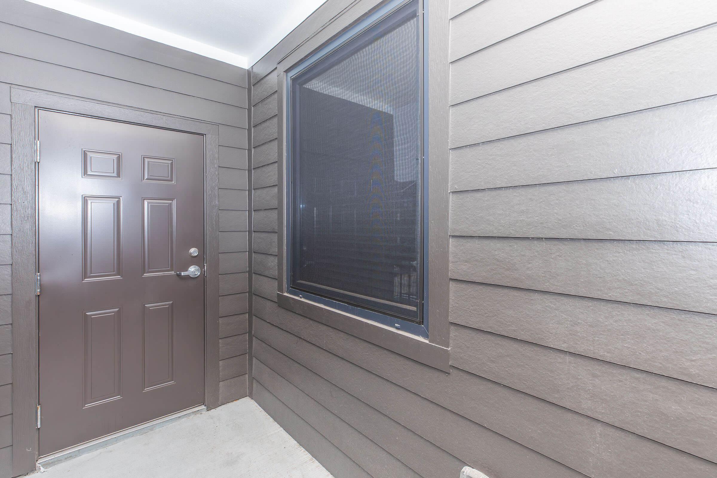 A modern exterior view of a brown wooden door with a sleek design, set against a wall featuring horizontal grooves. A large window beside the door reflects natural light, enhancing the entrance's contemporary appearance. The overall vibe is inviting and well-maintained.