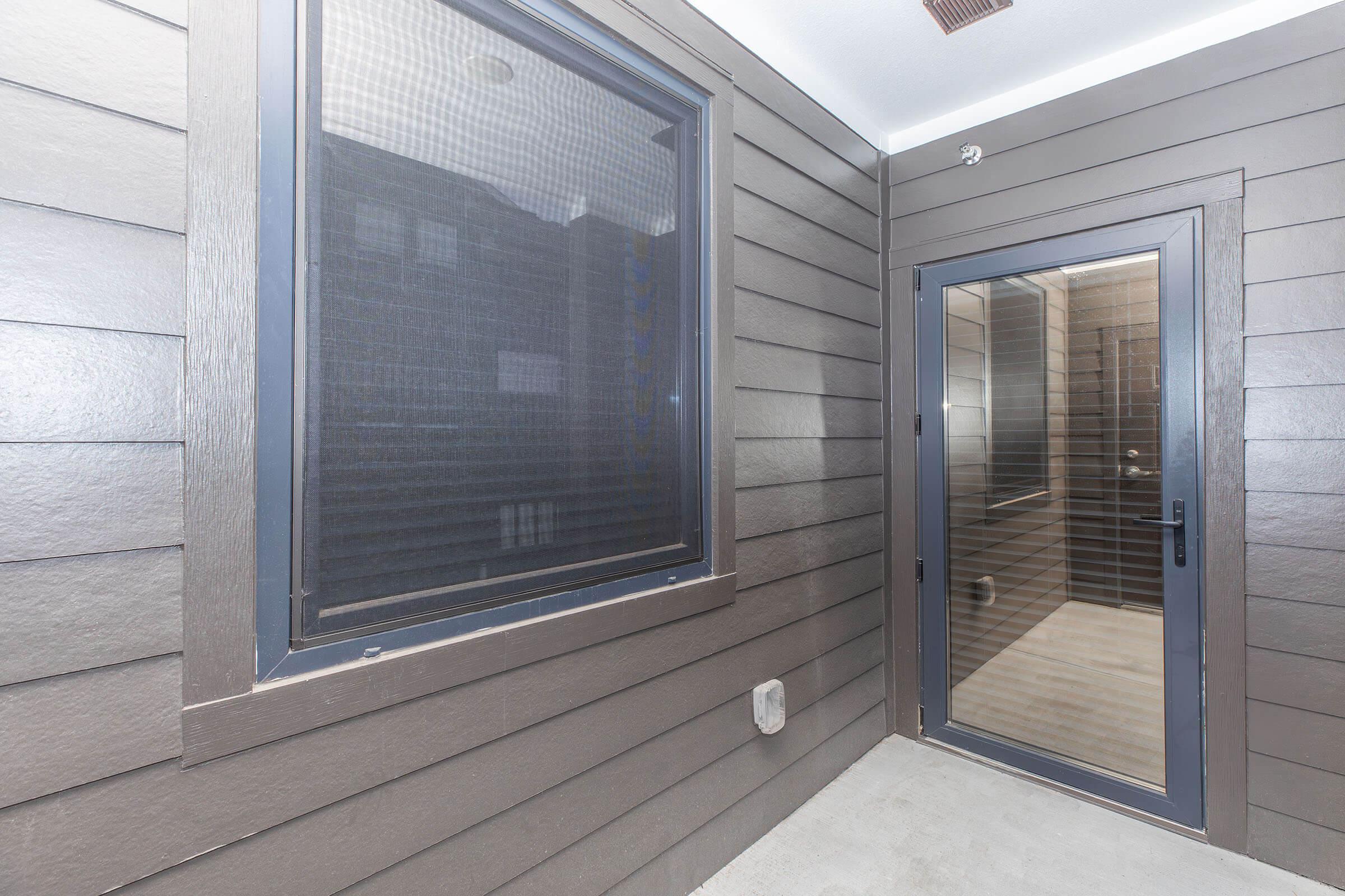 A modern entryway featuring a glass door and a large window with a screen. The walls are clad in dark wood siding, giving a sleek look. The space is well-lit, with light-colored flooring and minimal decor, creating an inviting and contemporary atmosphere.