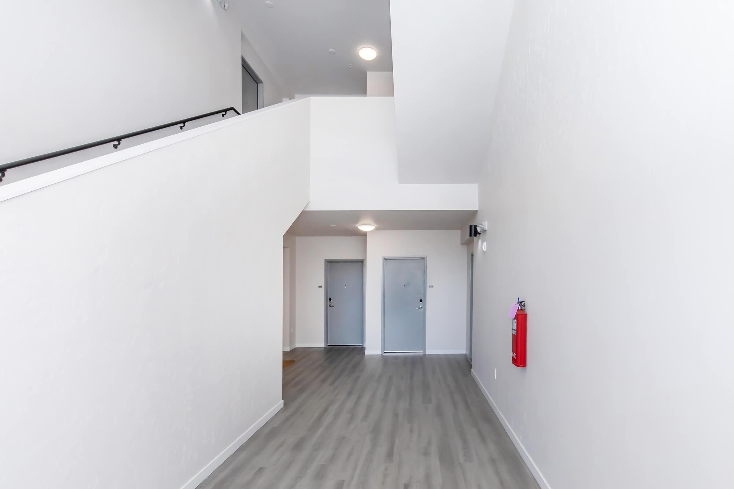 A bright, modern hallway featuring light-colored walls and a wooden floor. There are two closed doors on one side and a fire extinguisher mounted on the wall. The space is well-lit with natural light coming from above, creating an open and inviting atmosphere.
