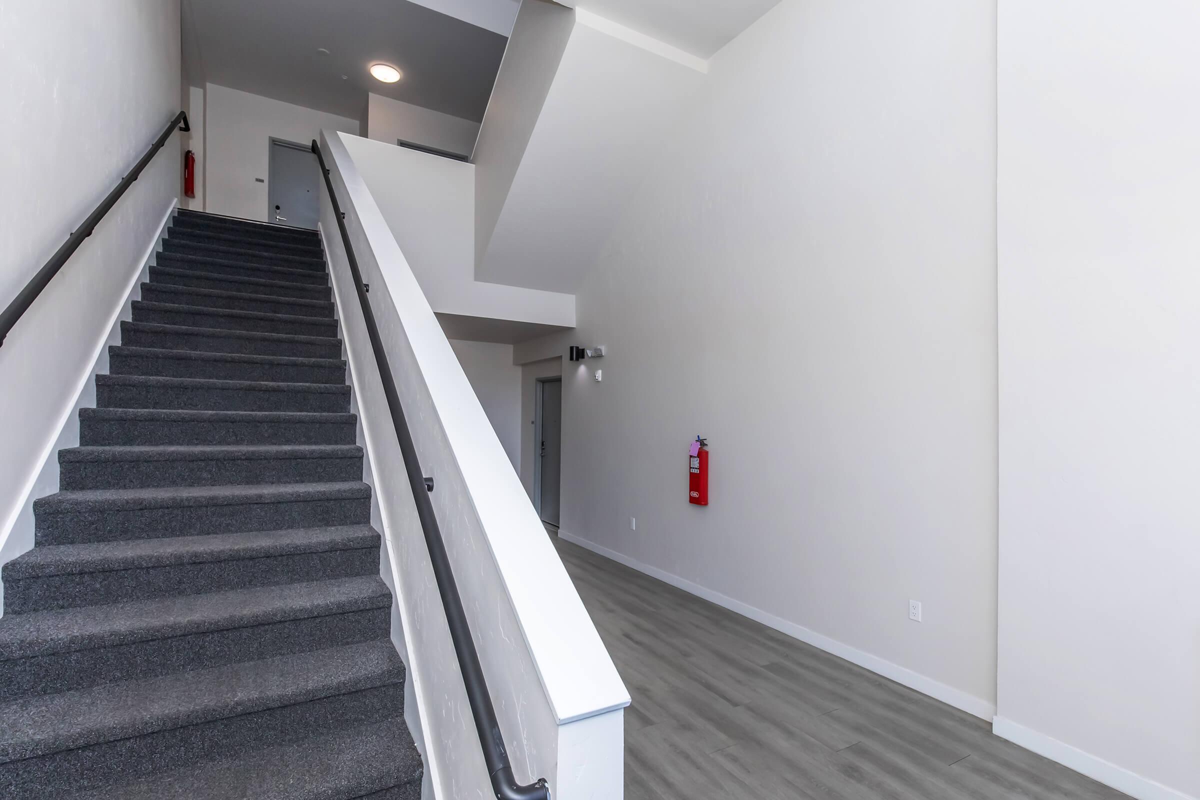 Interior view of a modern stairwell featuring gray carpeted stairs leading to an upper level. The walls are painted white and the floor is a light wood finish. A fire extinguisher is mounted on the wall to the right, and there is an absence of decorations or furniture, creating a clean and minimalist look.