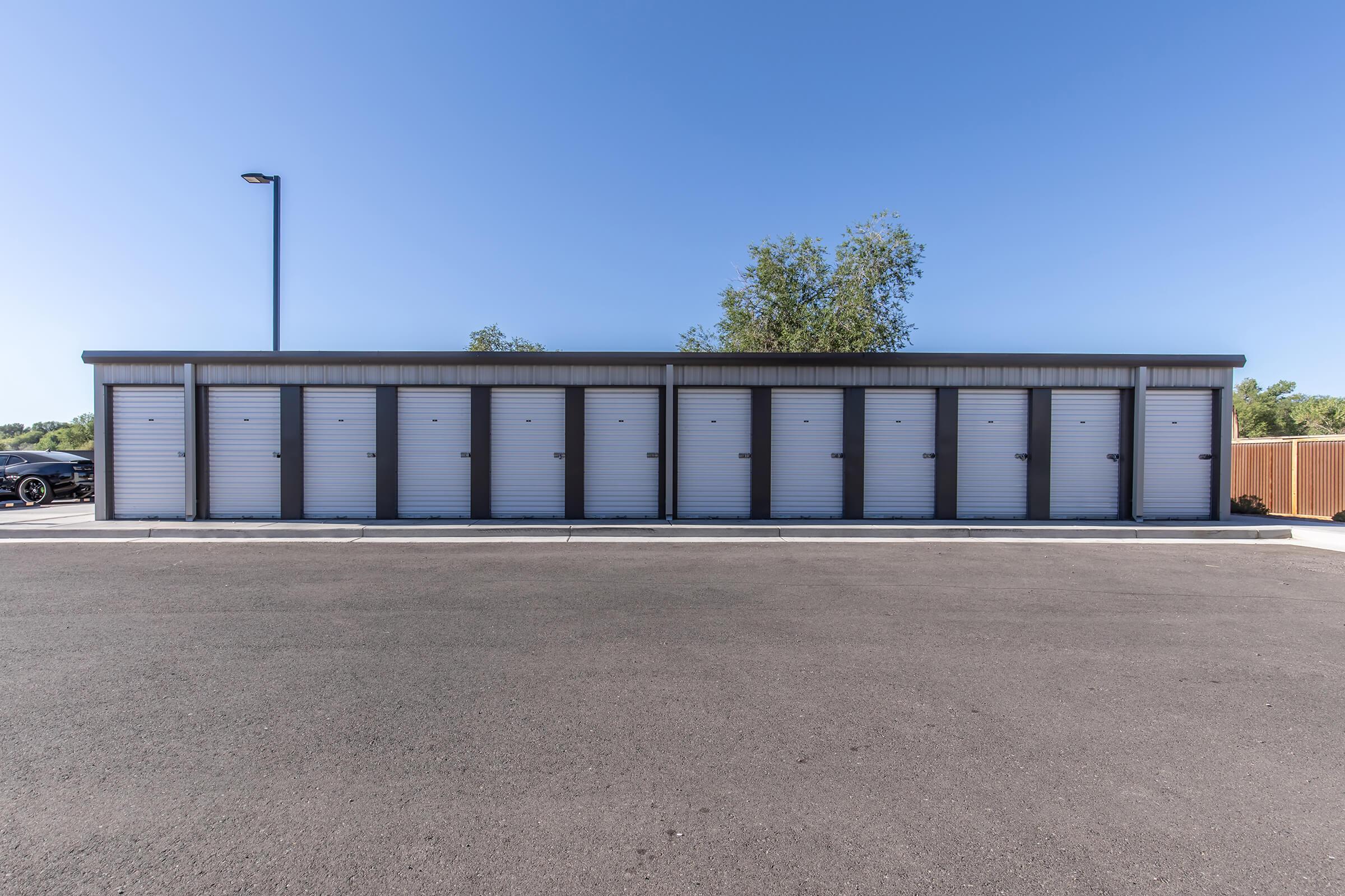 A row of storage units with individual roll-up doors, set against a clear blue sky. The units are situated on a paved lot, with a few trees in the background and a nearby parked vehicle.