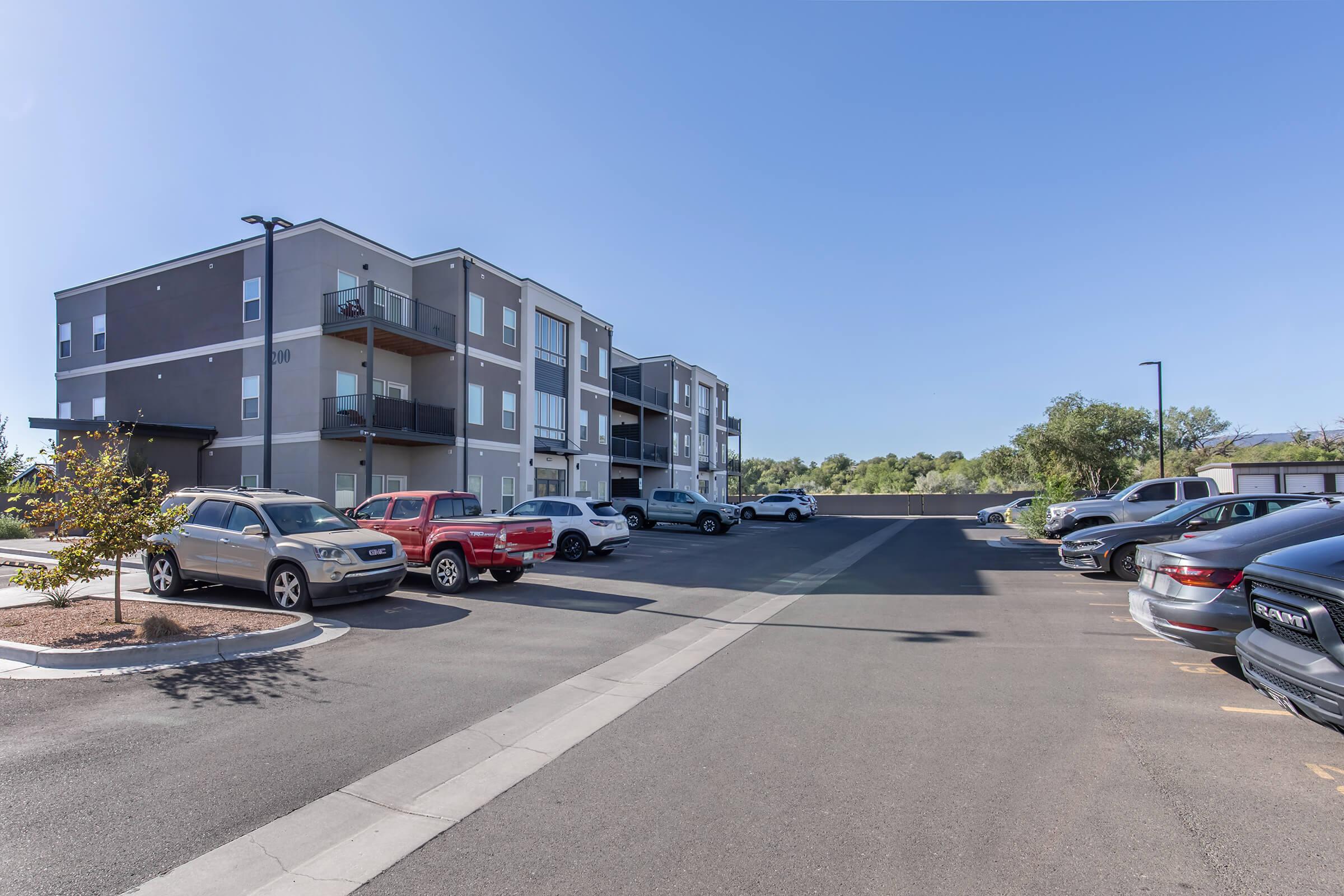 Modern apartment building with multiple stories and balconies, surrounded by parked cars in a paved lot. Clear blue sky overhead and trees in the background. The scene captures a typical residential area with a focus on convenience and accessibility.