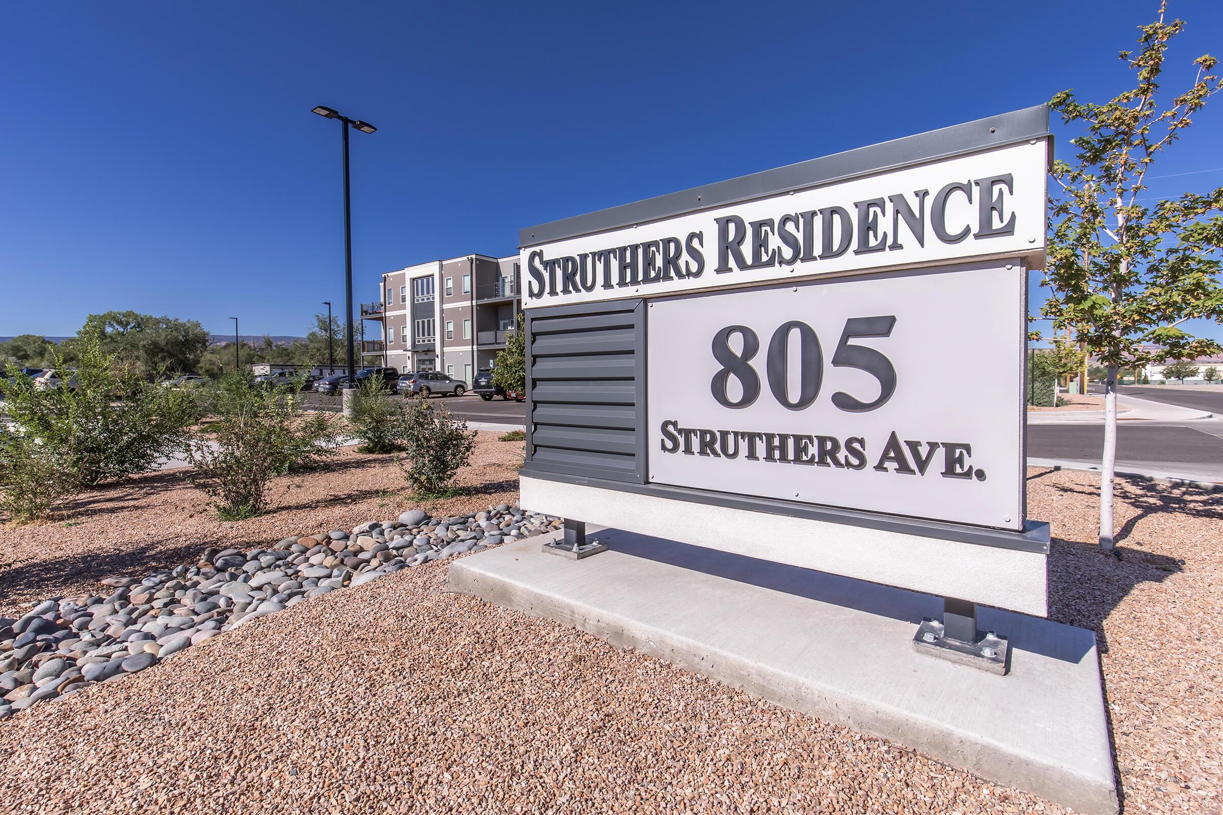 Sign for Struthers Residence located at 805 Struthers Ave. The sign features modern design elements and is surrounded by landscaped areas with small rocks and shrubs. In the background, there are multi-story residential buildings under a clear blue sky.