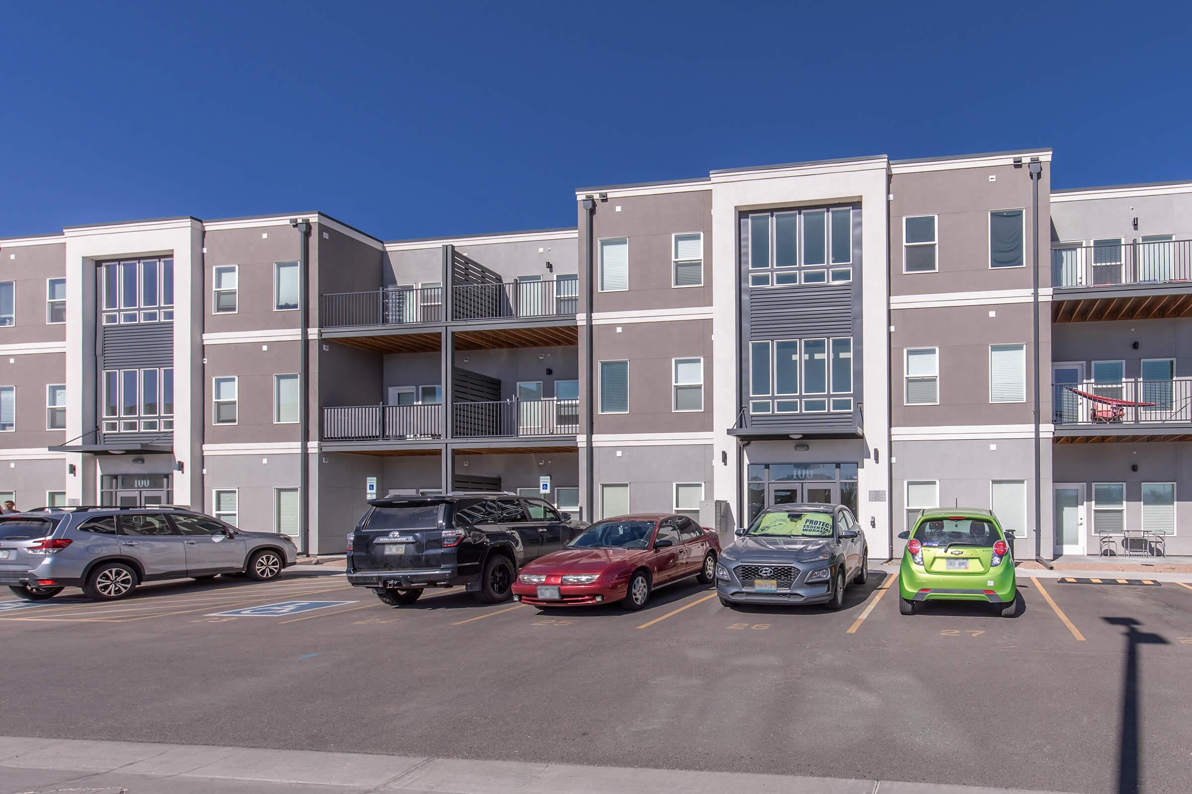 A modern multi-unit apartment building with a gray exterior and several balconies. The front features a parking area with various cars, including a green vehicle. Clear blue sky above and structured landscaping around the building.