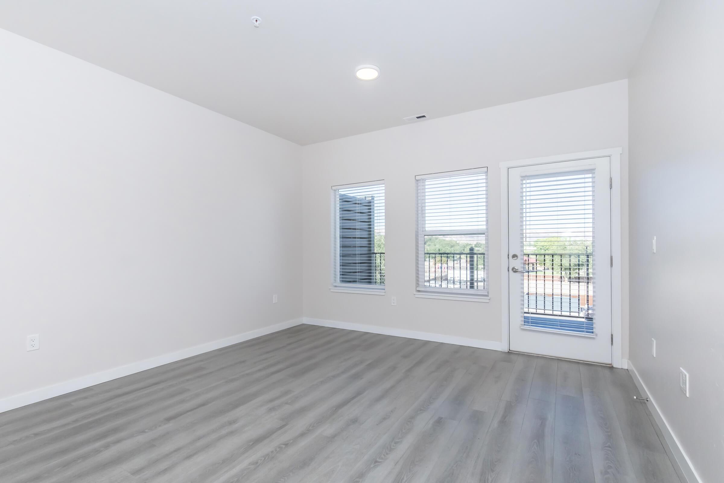Spacious and well-lit room featuring light gray walls, large windows with blinds, and a sliding glass door leading to a balcony. The flooring is light wood-like laminate, providing a modern and clean look, with minimal furnishings for a versatile space.